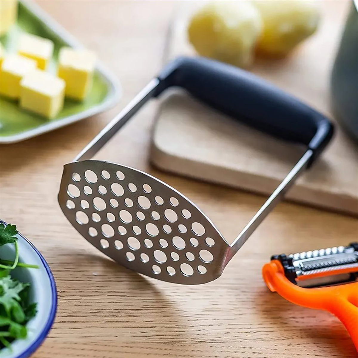 Kartoffelstampfer, Butter und ein Schäler auf einer Holzoberfläche.