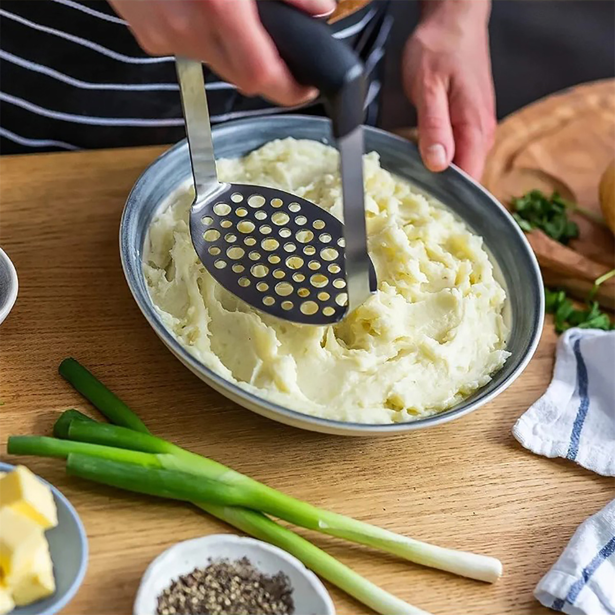 Eine Person zerdrückt Kartoffeln in einer Schüssel. Grüne Zwiebeln und Butter sind in der Nähe.