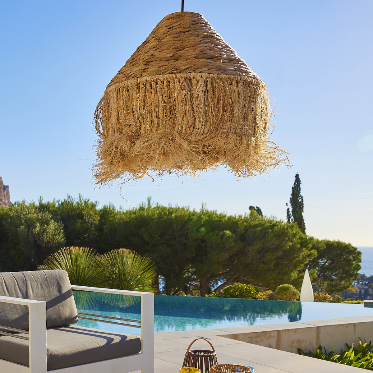 Eine gewebte Strohlampe hängt über einem Pool. Bäume und Himmel bilden den Hintergrund.
