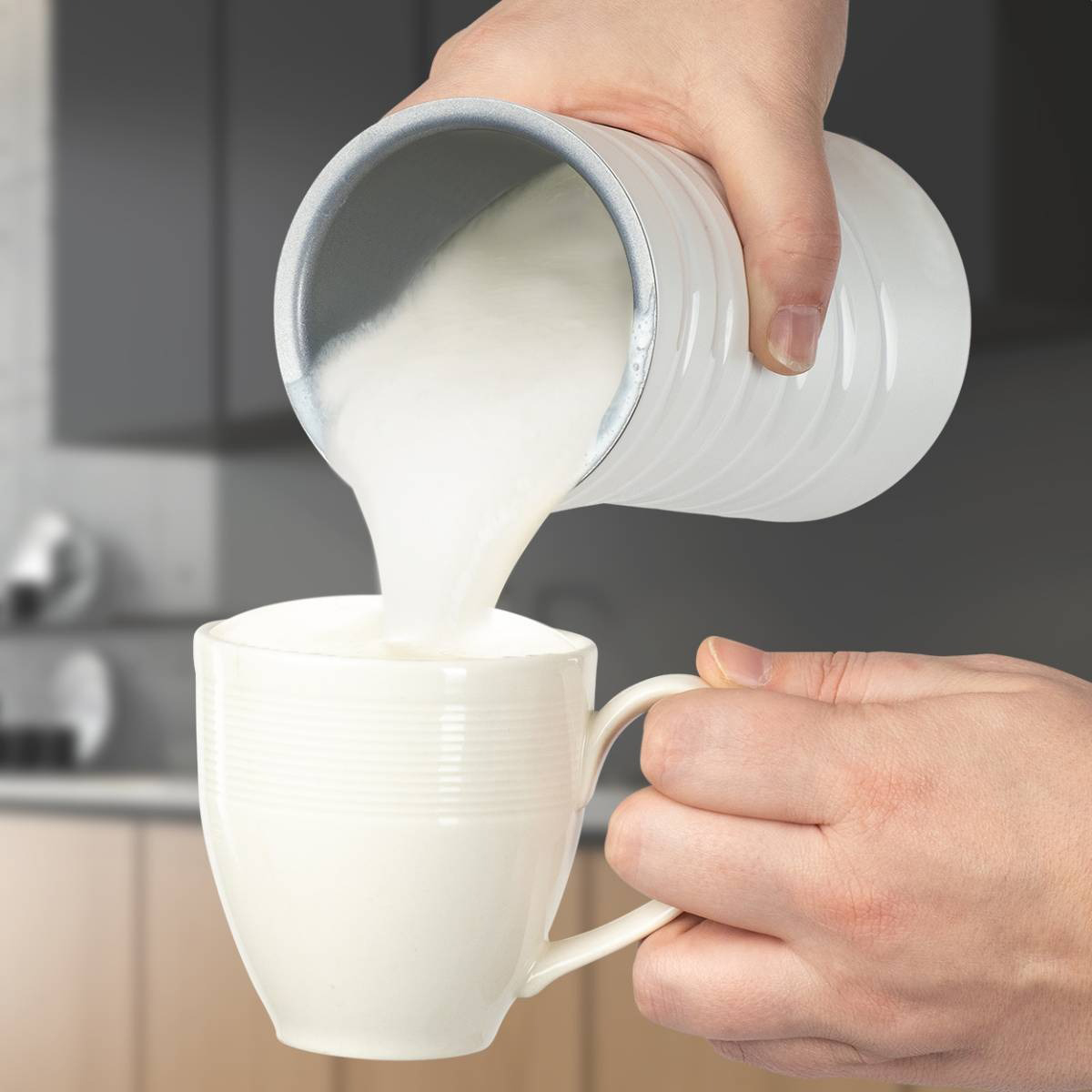 Hände gießen Milch aus einem weißen Krug in eine weiße Tasse, neutraler Hintergrund.