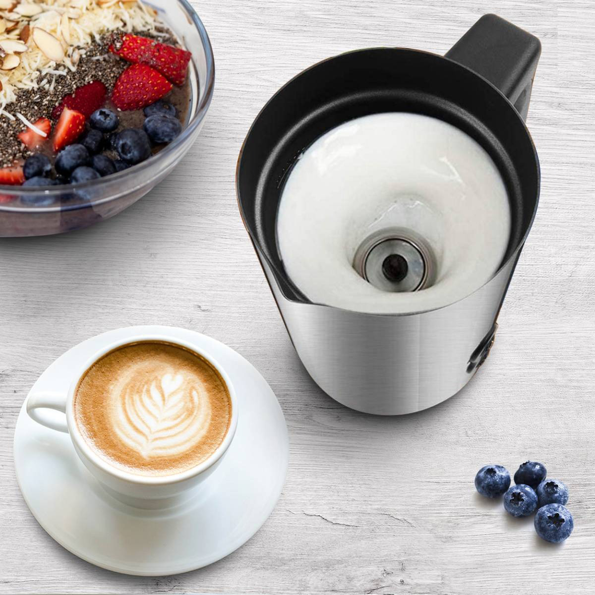 Eine Tasse Kaffee mit Latte Art neben einer Schüssel mit Obst und dem Milchaufschäumer.