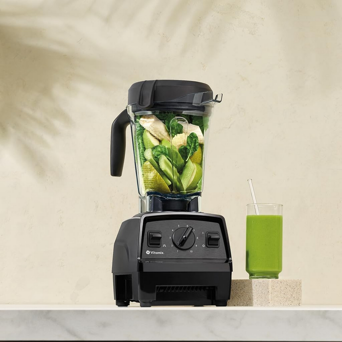 Schwarzer Mixer mit Zutaten, Glas grünen Smoothies auf weißer Oberfläche.