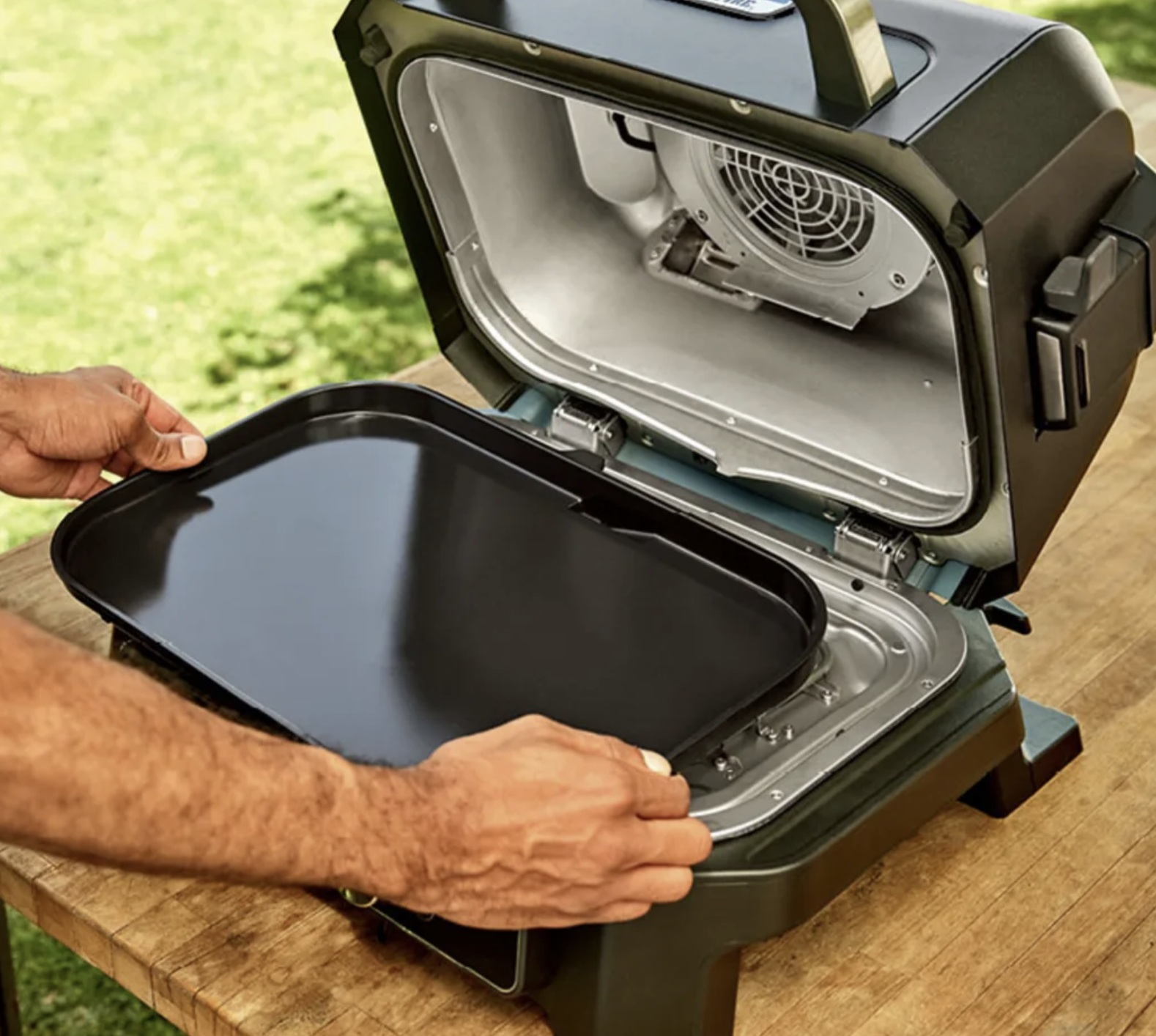 Ręce trzymające czarną tacę do grilla, otwarty przenośny grill. Wnętrze jest srebrne i czarne.