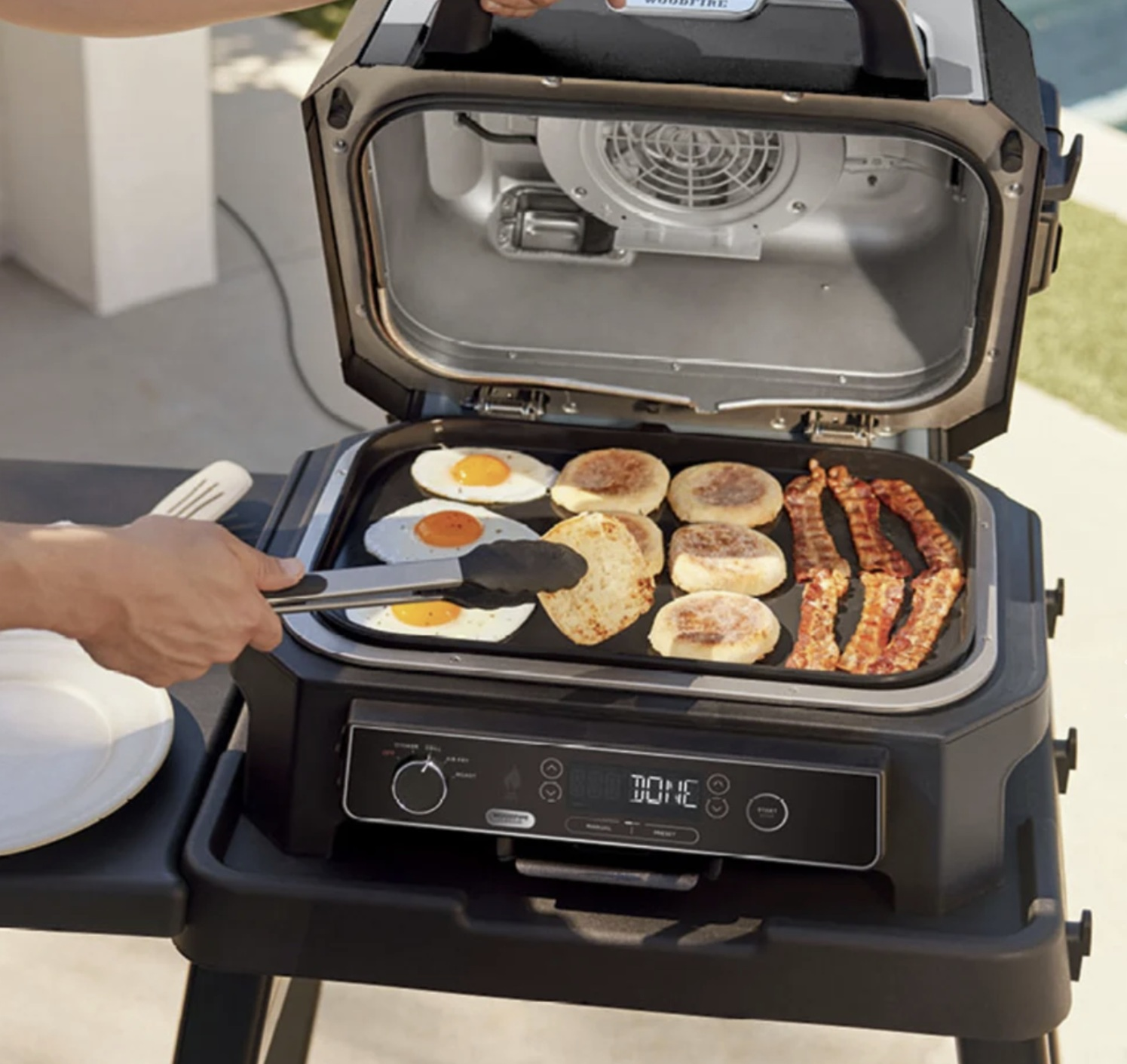 Urządzenie do gotowania na zewnątrz z jedzeniem, jajkami, naleśnikami i boczkiem. Ręka grilluje jedzenie.