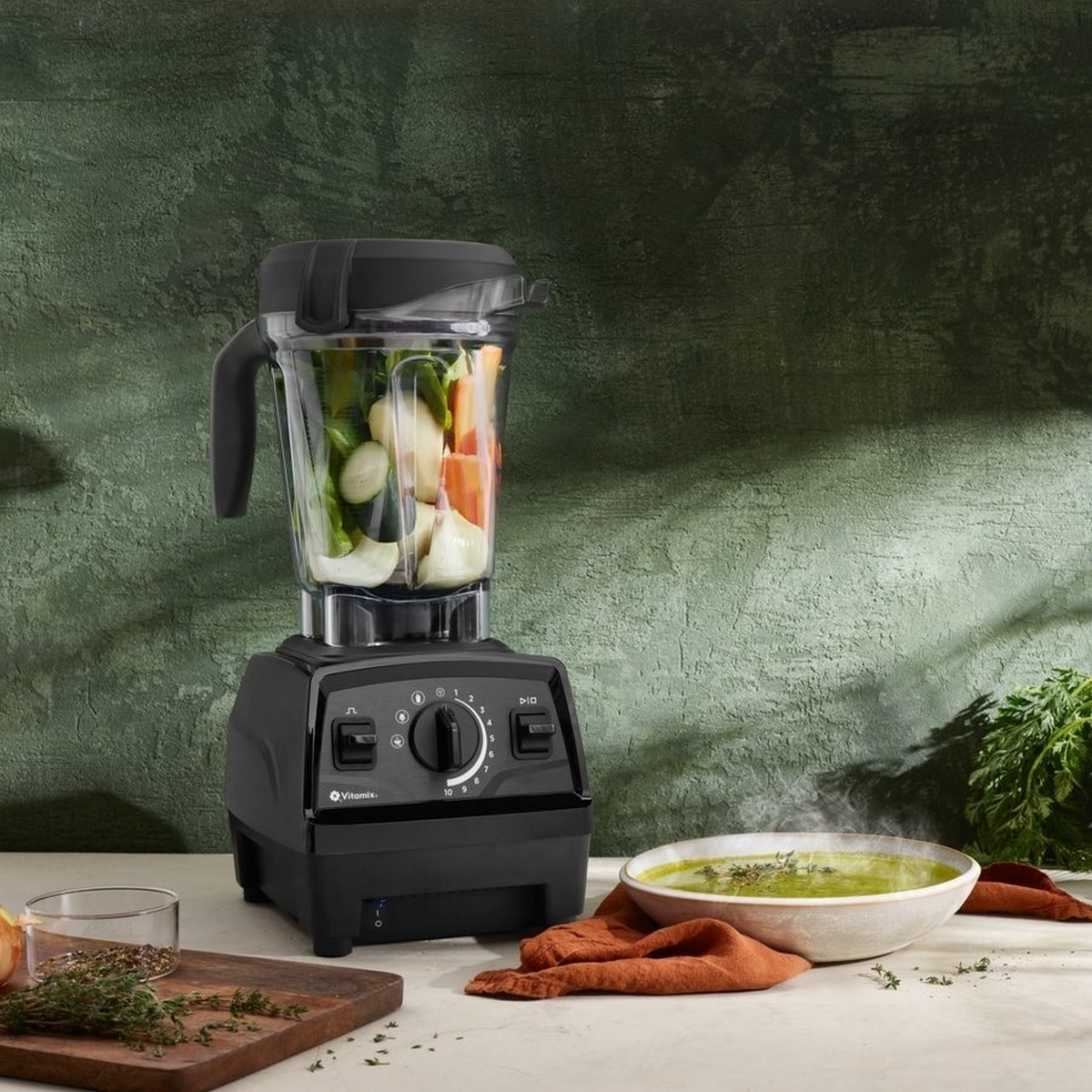 Schwarzer Vitamix-Mixer mit Zutaten, neben einer Suppenschüssel und einem Holzbrett.