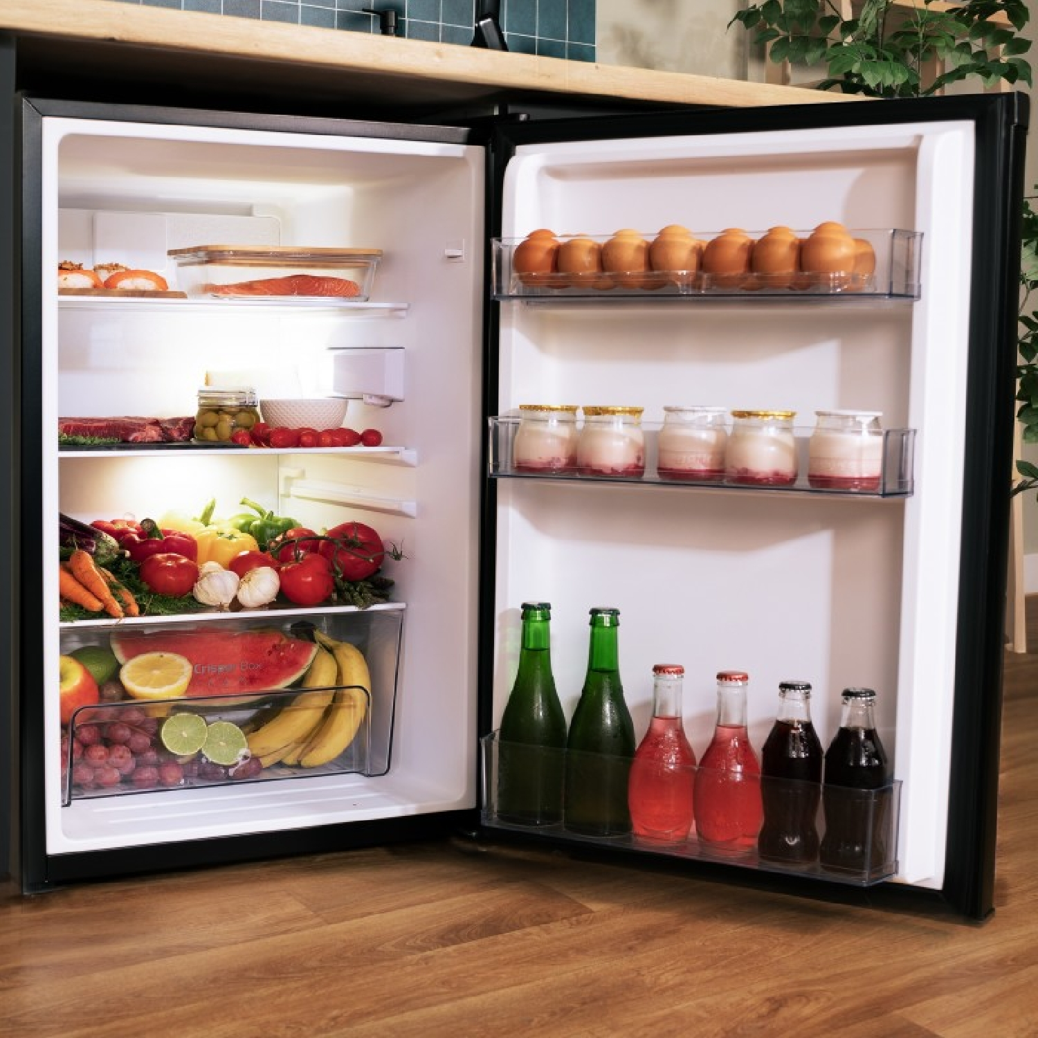 Refrigerador abierto con comida. Huevos, yogur, botellas, verduras, fruta. Suelo de madera.