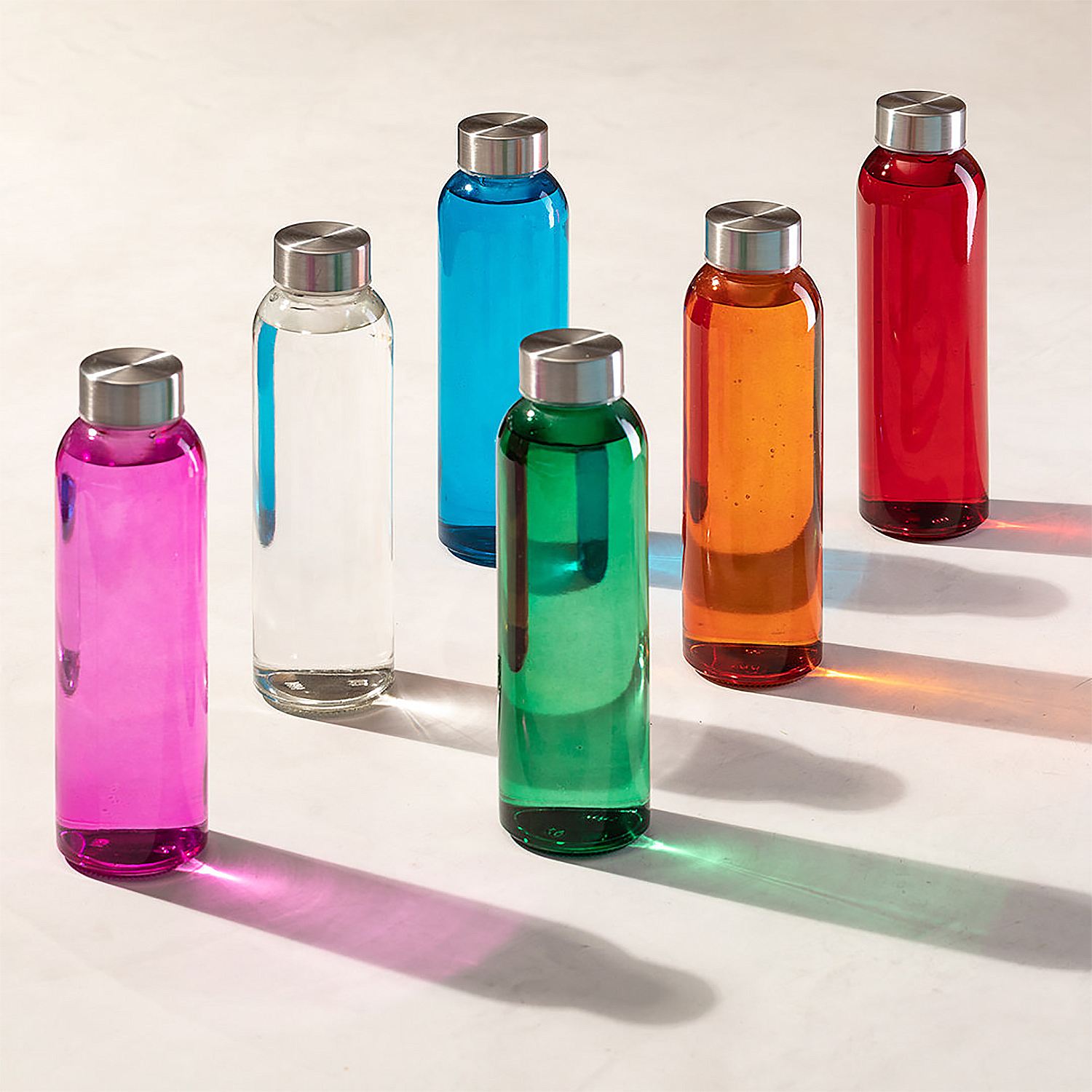 Botellas de agua coloridas con líquidos de diferentes colores. Tapas plateadas, superficie blanca.