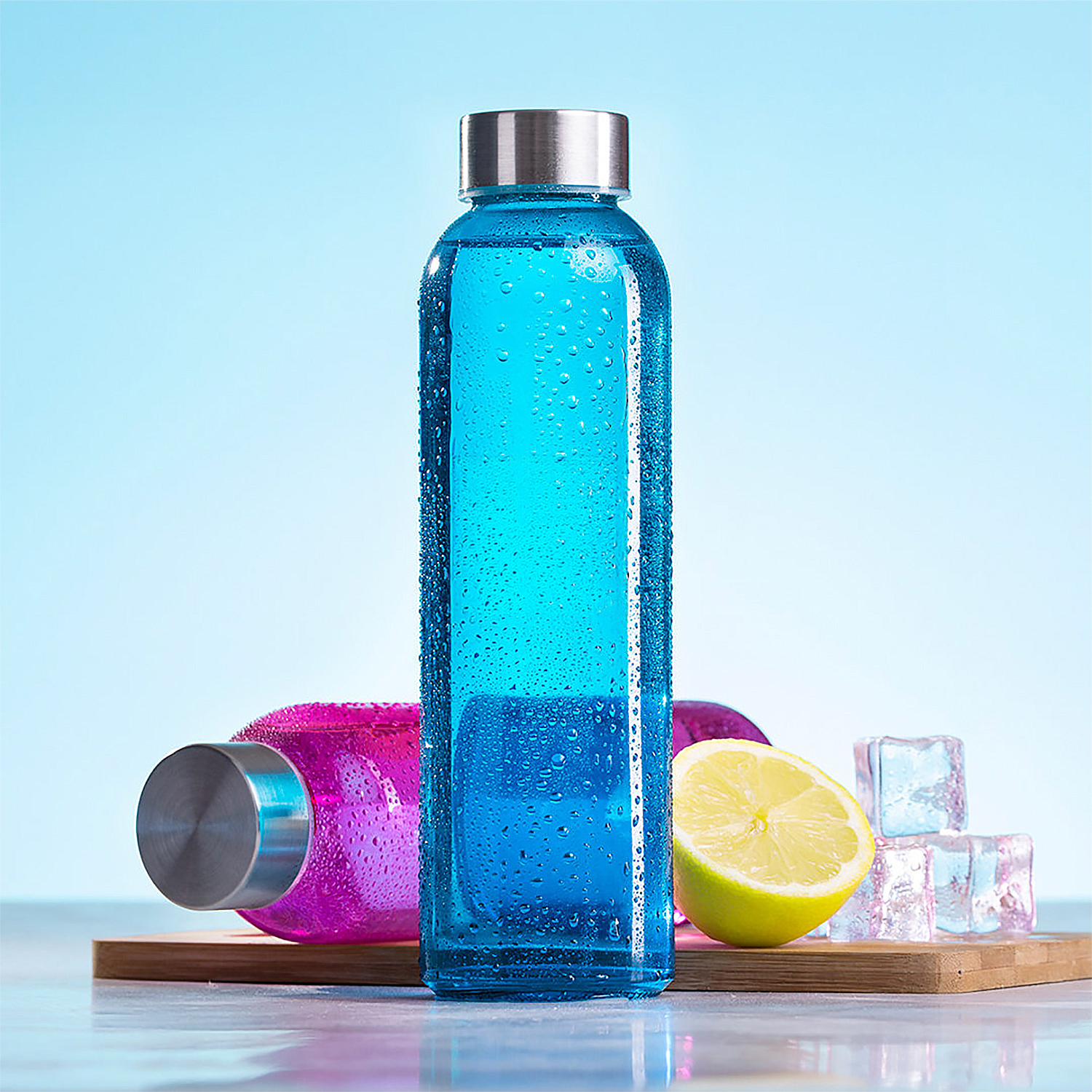 Botella de agua azul, botella rosa, mitad de limón y cubitos de hielo en tabla de madera, fondo azul.