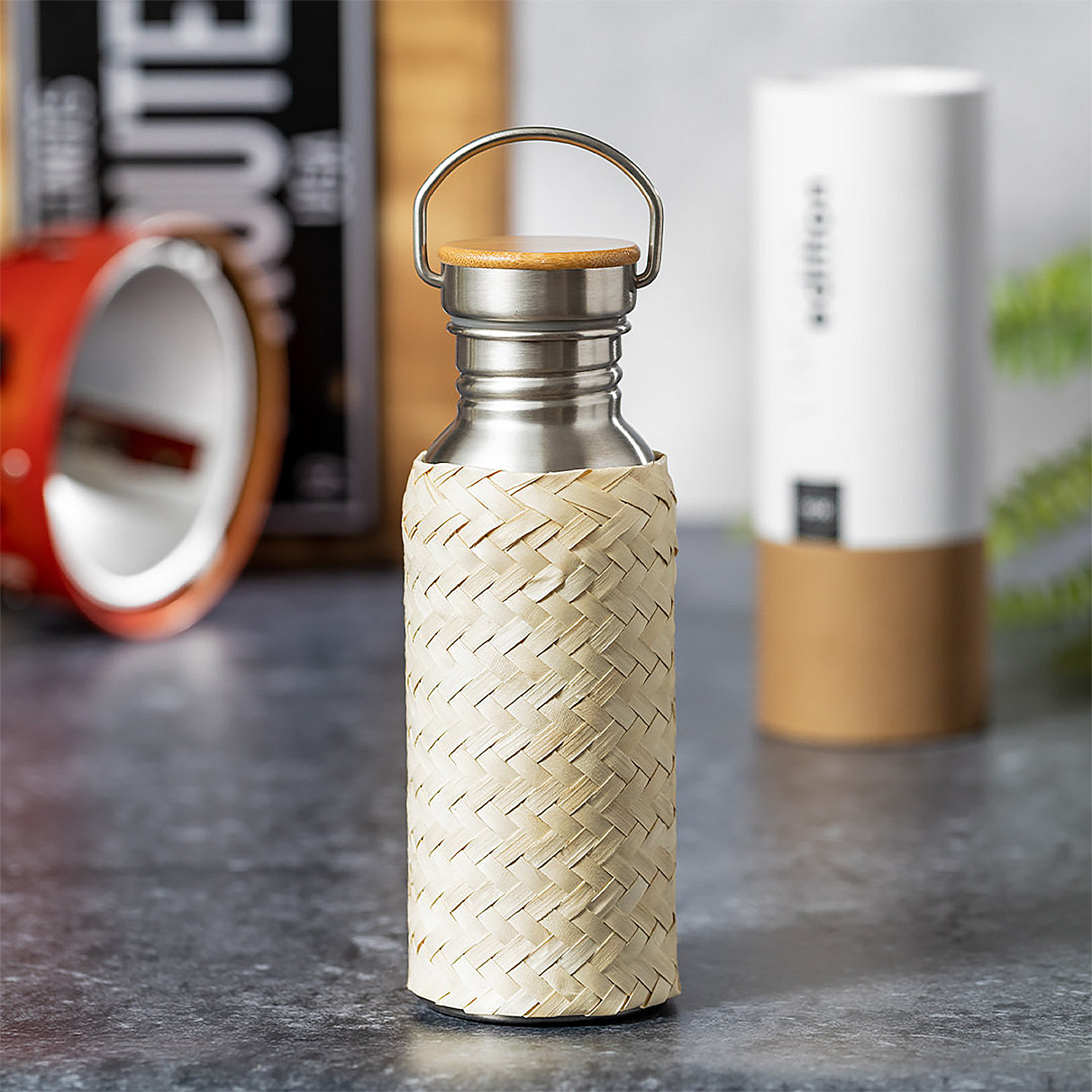 Una botella de agua de acero inoxidable envuelta en bambú tejido. Un recipiente blanco al fondo.