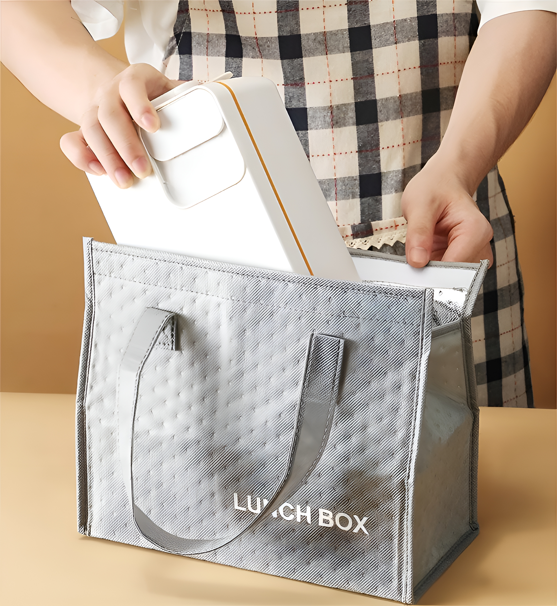 Eine Person legt eine weiße Lunchbox in eine silberne isolierte Lunchtasche. Eine karierte Schürze wird getragen.