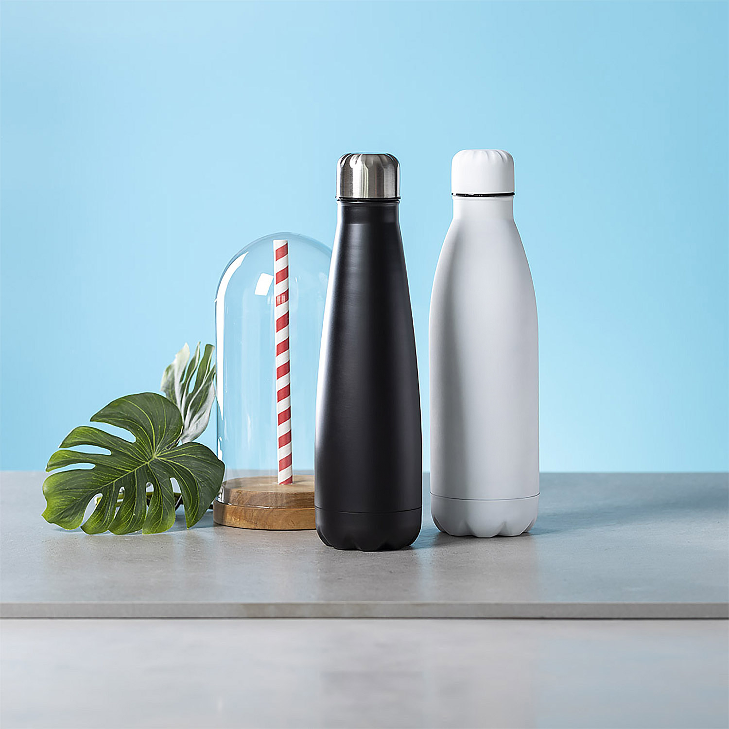 Dos botellas de agua, una negra y otra blanca, sobre una superficie, una pajita bajo una cúpula de cristal al fondo.