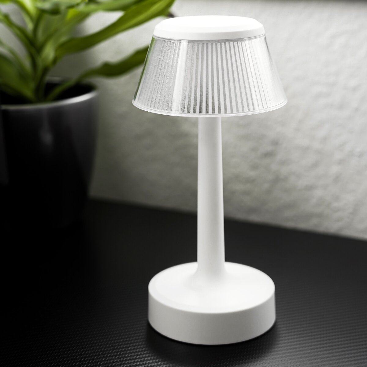 Weiße Tischlampe. Zylindrischer Sockel, Schaft und strukturierter Schirm. Schwarzer Tisch, grüner Hintergrund.