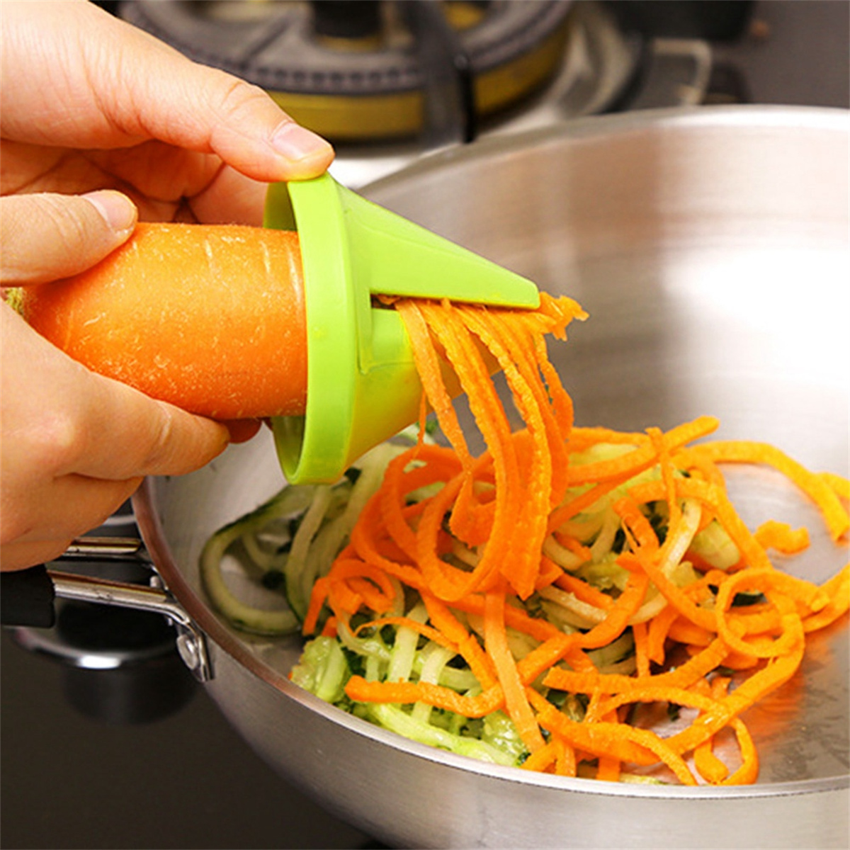 Eine Hand verwendet einen Spiralschneider, um eine Karotte in eine Pfanne mit anderen Karotten- und Zucchinispiralen zu raspeln.