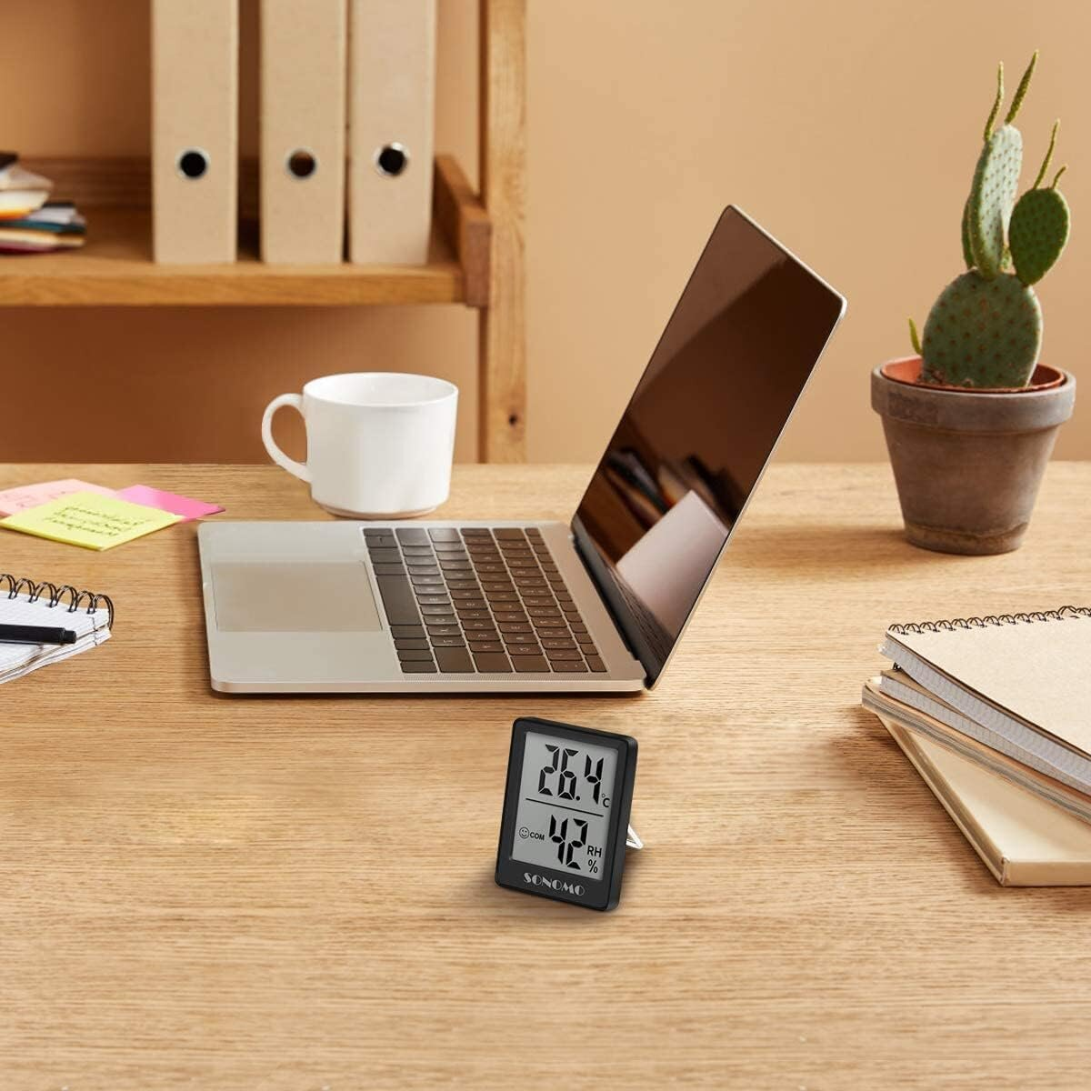 Ein digitales Thermometer auf einem Schreibtisch mit Laptop, Kaffeetasse und einigen Notizbüchern. Es zeigt 26,4 Grad an.