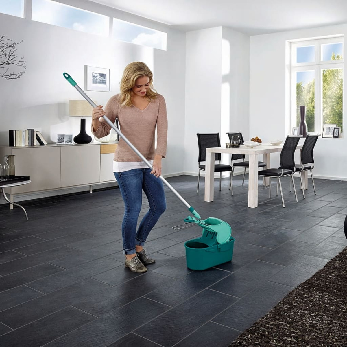 Une femme lave un sol sombre avec une serpillère et un seau verts dans un salon moderne.