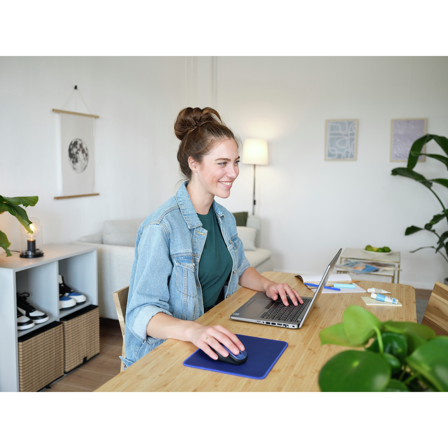 Kobieta używa laptopa przy drewnianym biurku z niebieską podkładką, uśmiechnięta.