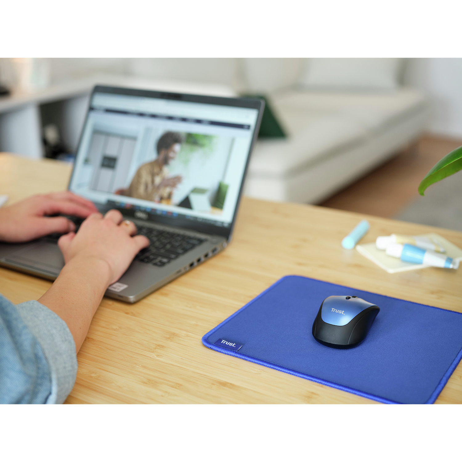 Osoba pisząca na laptopie, niebieska podkładka pod mysz i bezprzewodowa mysz na drewnianym stole. Tło jest rozmyte.