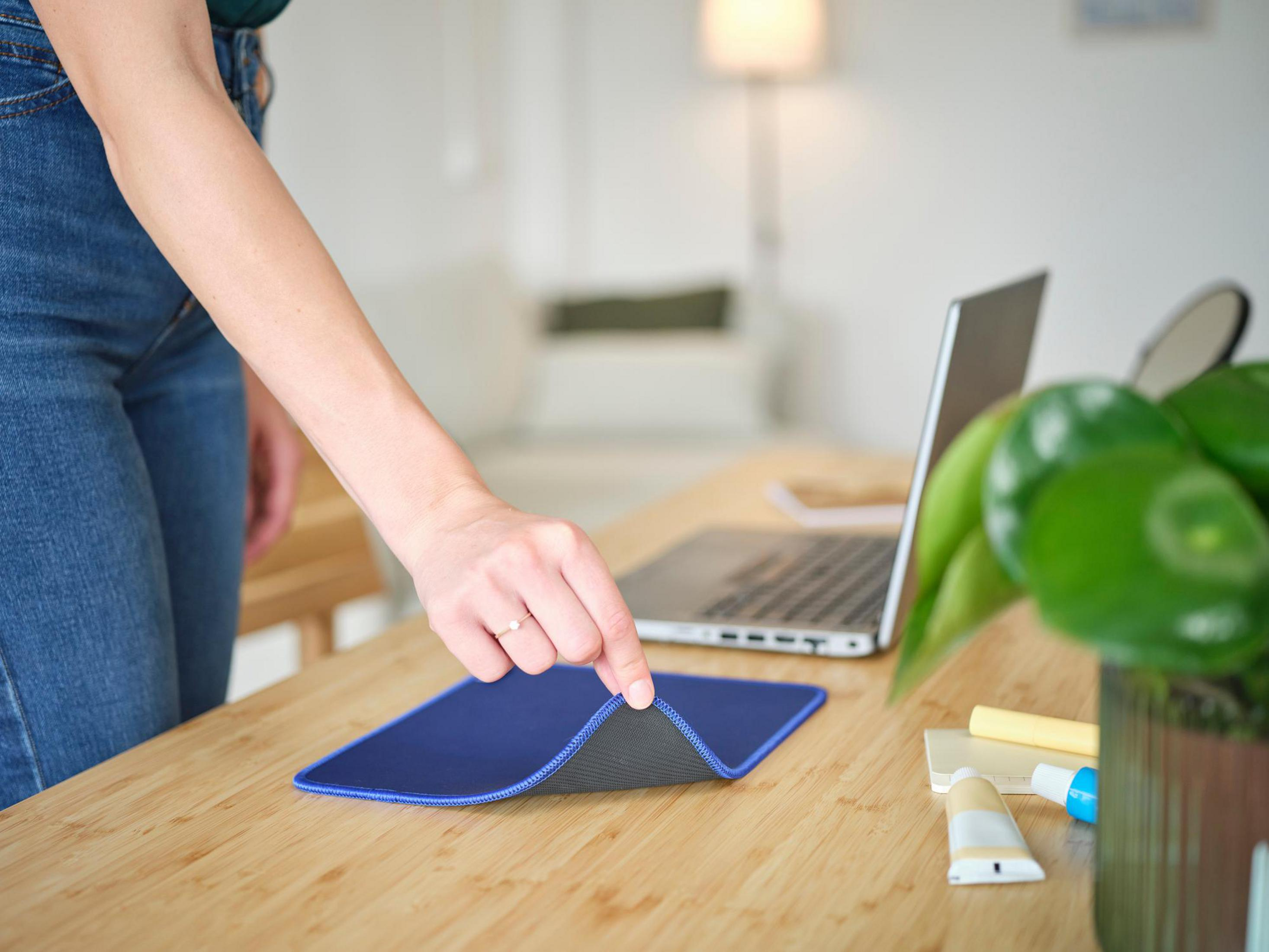Ręka osoby umieszczająca niebieską podkładkę pod mysz na drewnianym biurku. Na biurku znajduje się również laptop i roślina.