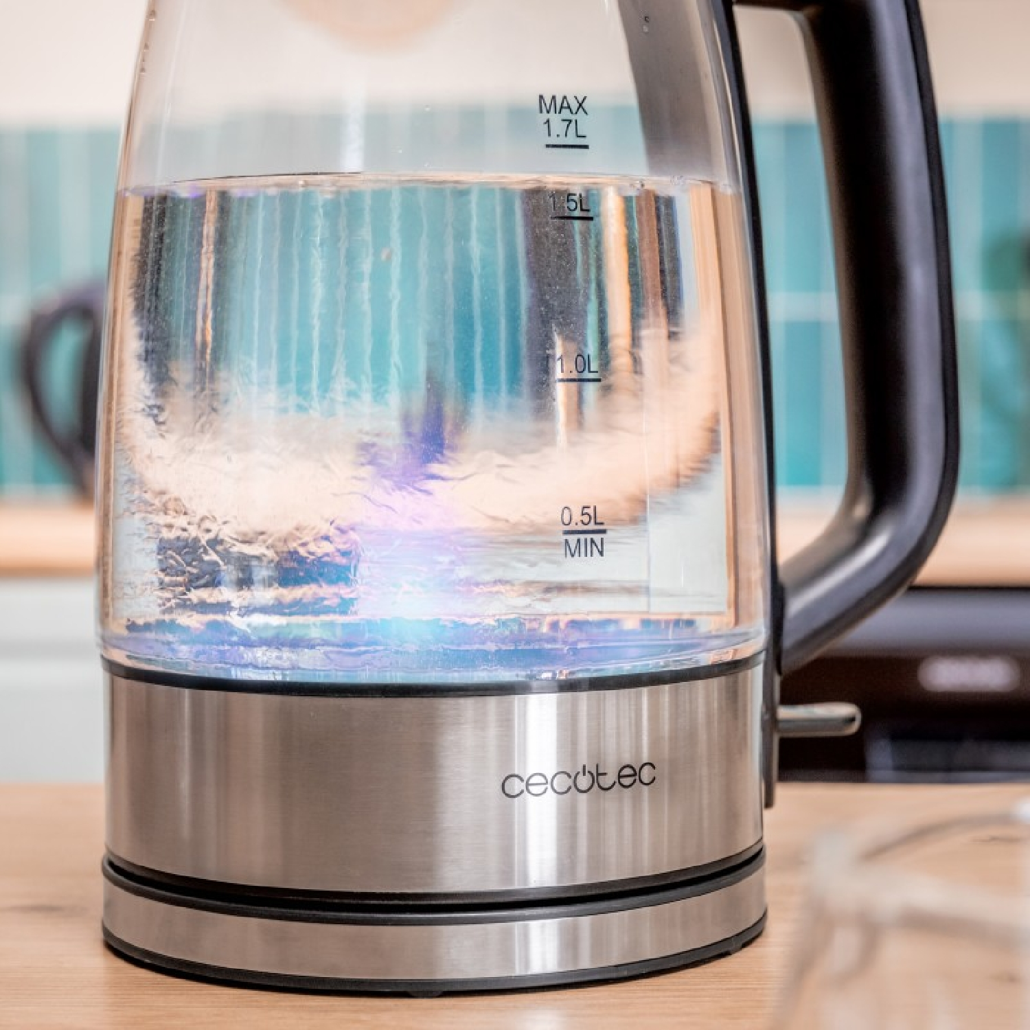 Een glazen elektrische waterkoker met water erin. Het heeft een zwarte handgreep en een zilveren basis, tegen een keukenachtergrond.