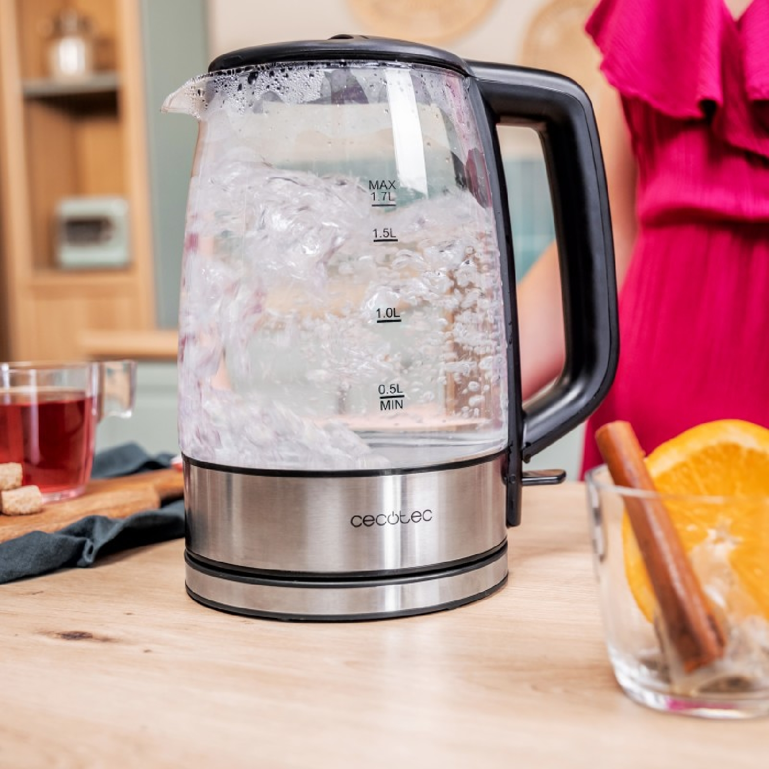 Glazen elektrische waterkoker met kokend water, een vrouw en ingrediënten op een houten tafel.