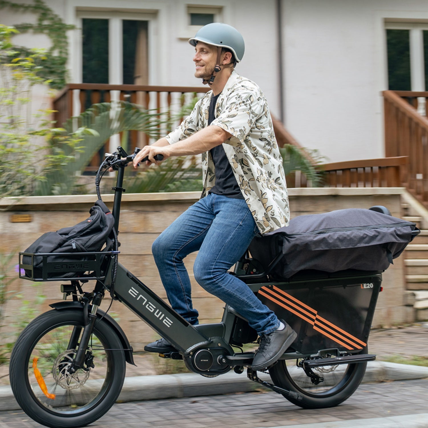 Uomo in bicicletta elettrica per strada. Indossa un casco e abiti casual. È presente un box nero.