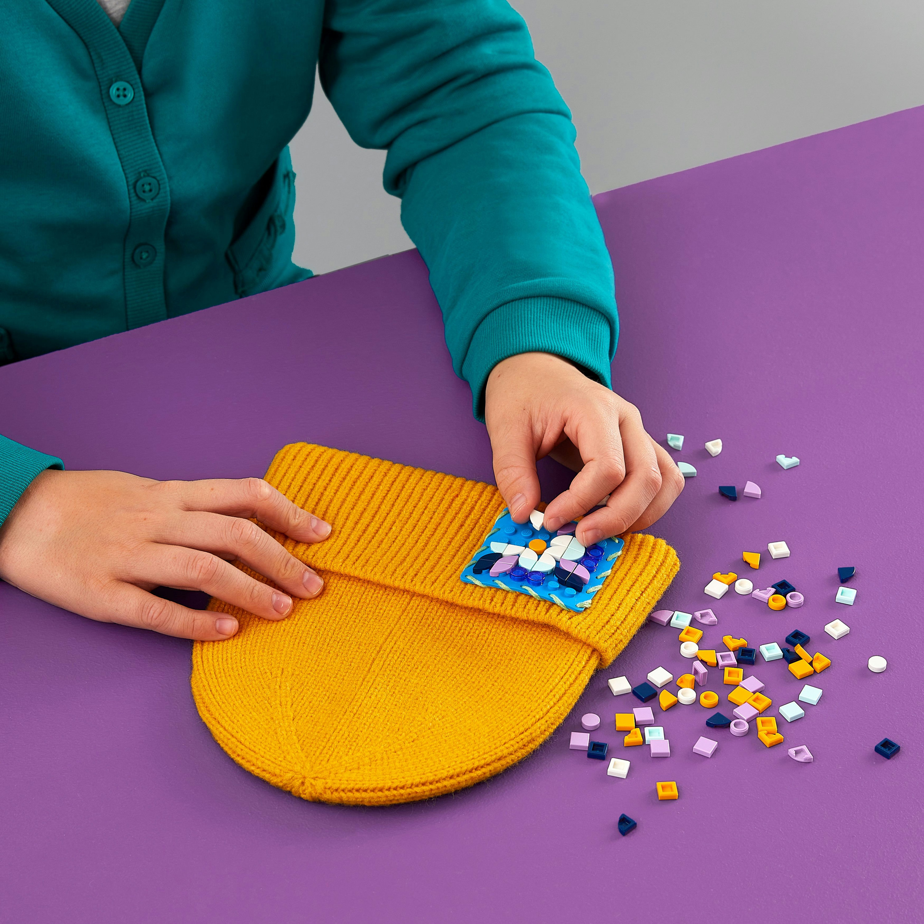 Ręce na fioletowym stole, szydełkujące żółtą czapkę z kolorowymi kwadratowymi elementami.