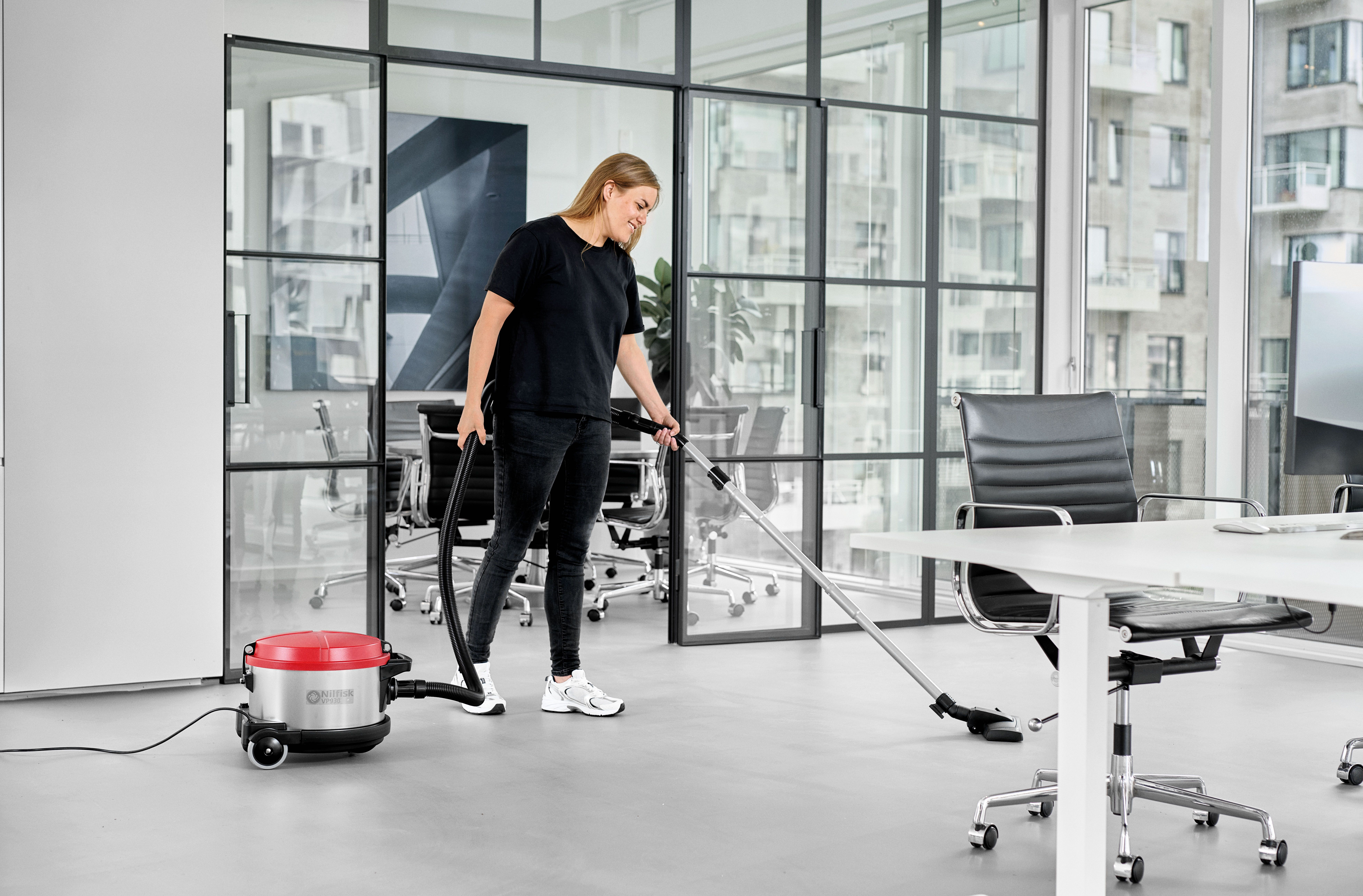 Kobieta odkurza biuro. Czerwony odkurzacz, szklane ściany, białe stoły.