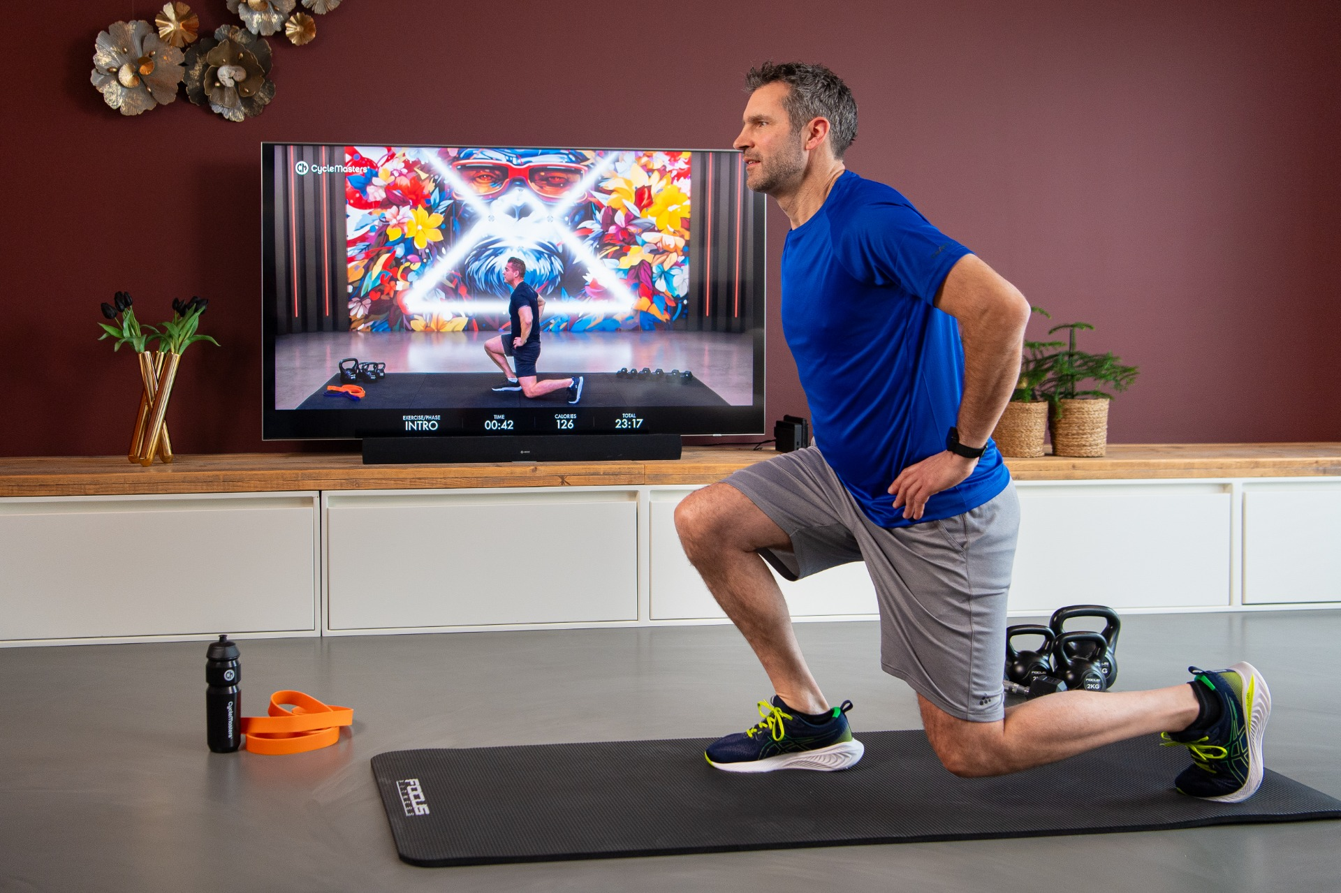 Un homme faisant des fentes avec des poids. Il est dans une salle de sport à domicile avec une télé montrant l'entraînement.