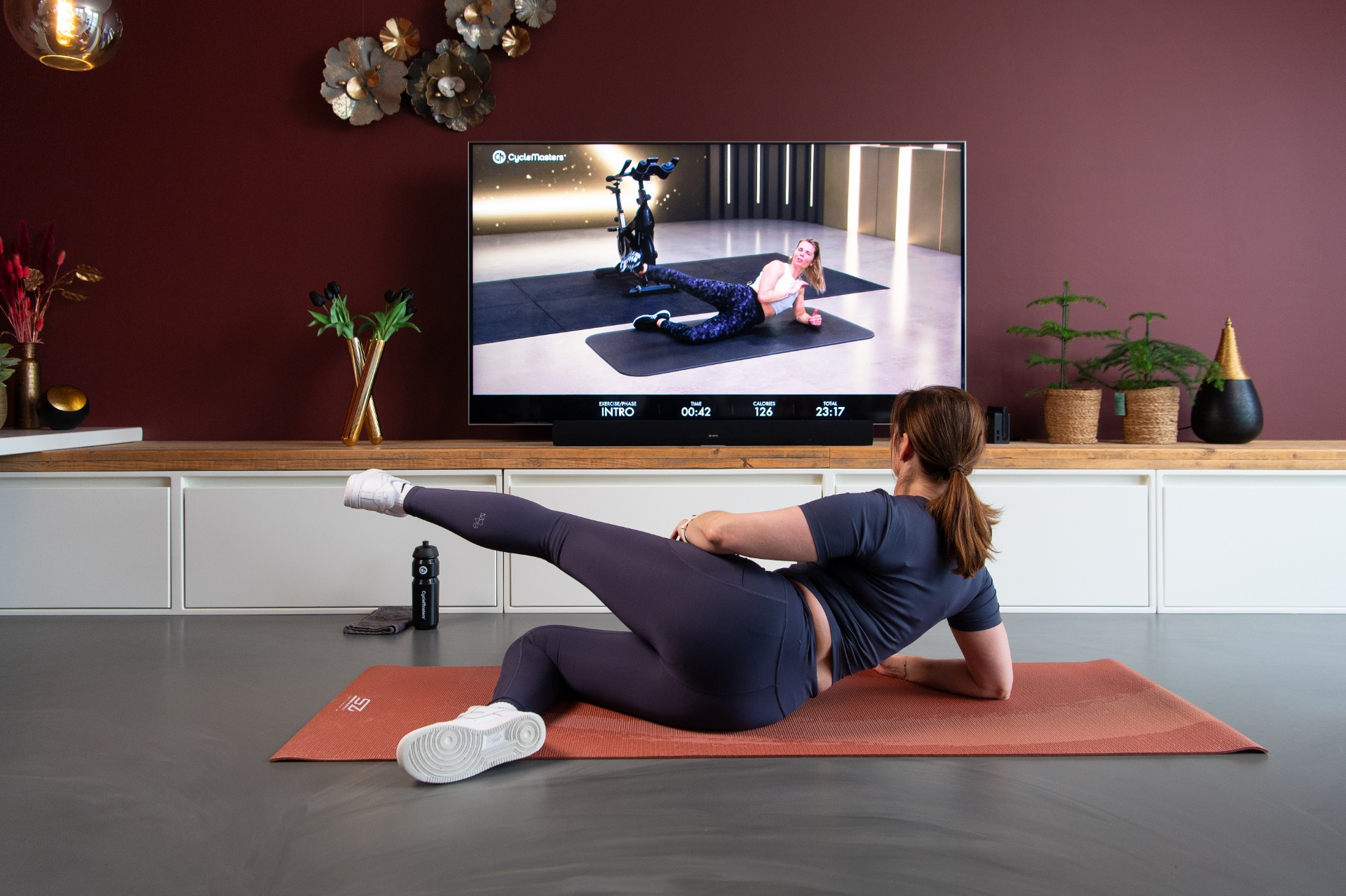 Woman exercising at home, watching a TV with exercise video. Red mat and plants.