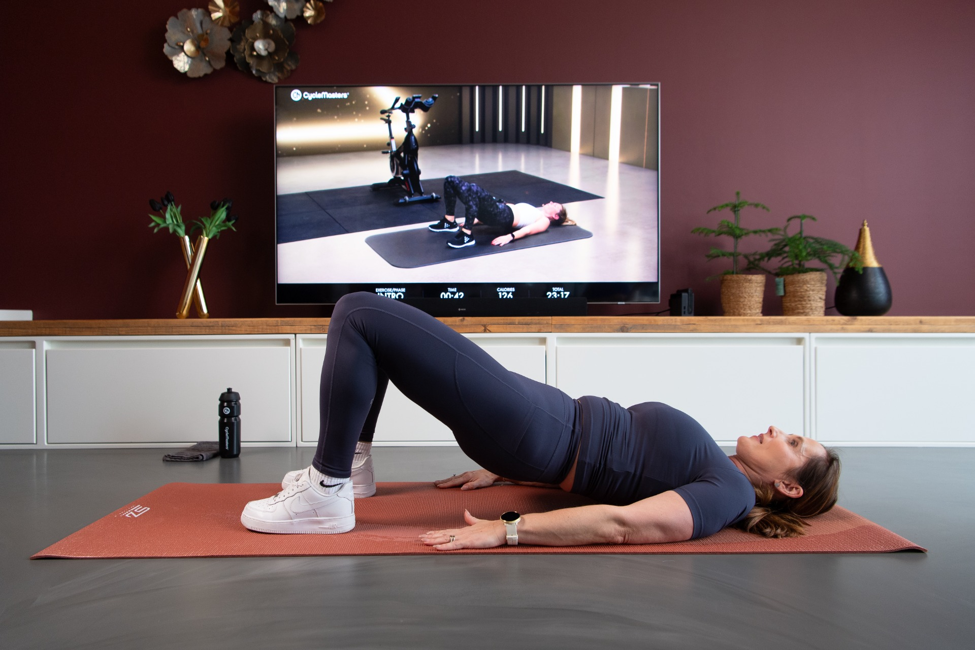 Woman doing bridge exercise on a mat in front of a TV screen.