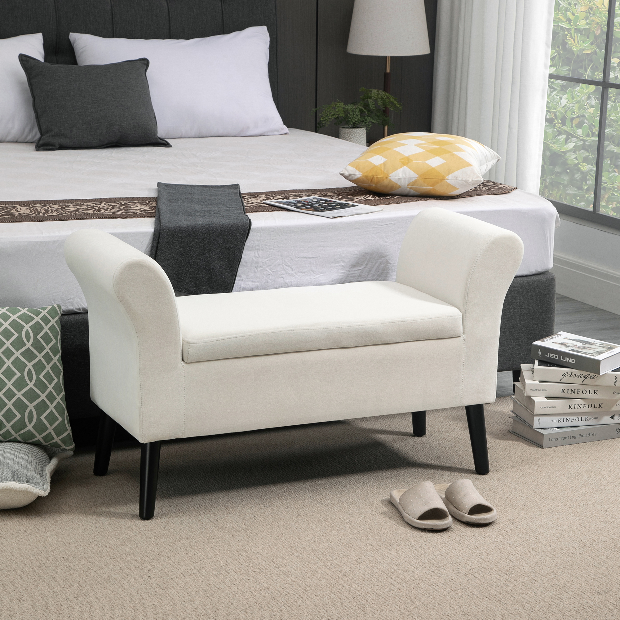 Banqueta blanquecina con brazos curvos y patas negras en un dormitorio, cerca de la cama y la ventana.