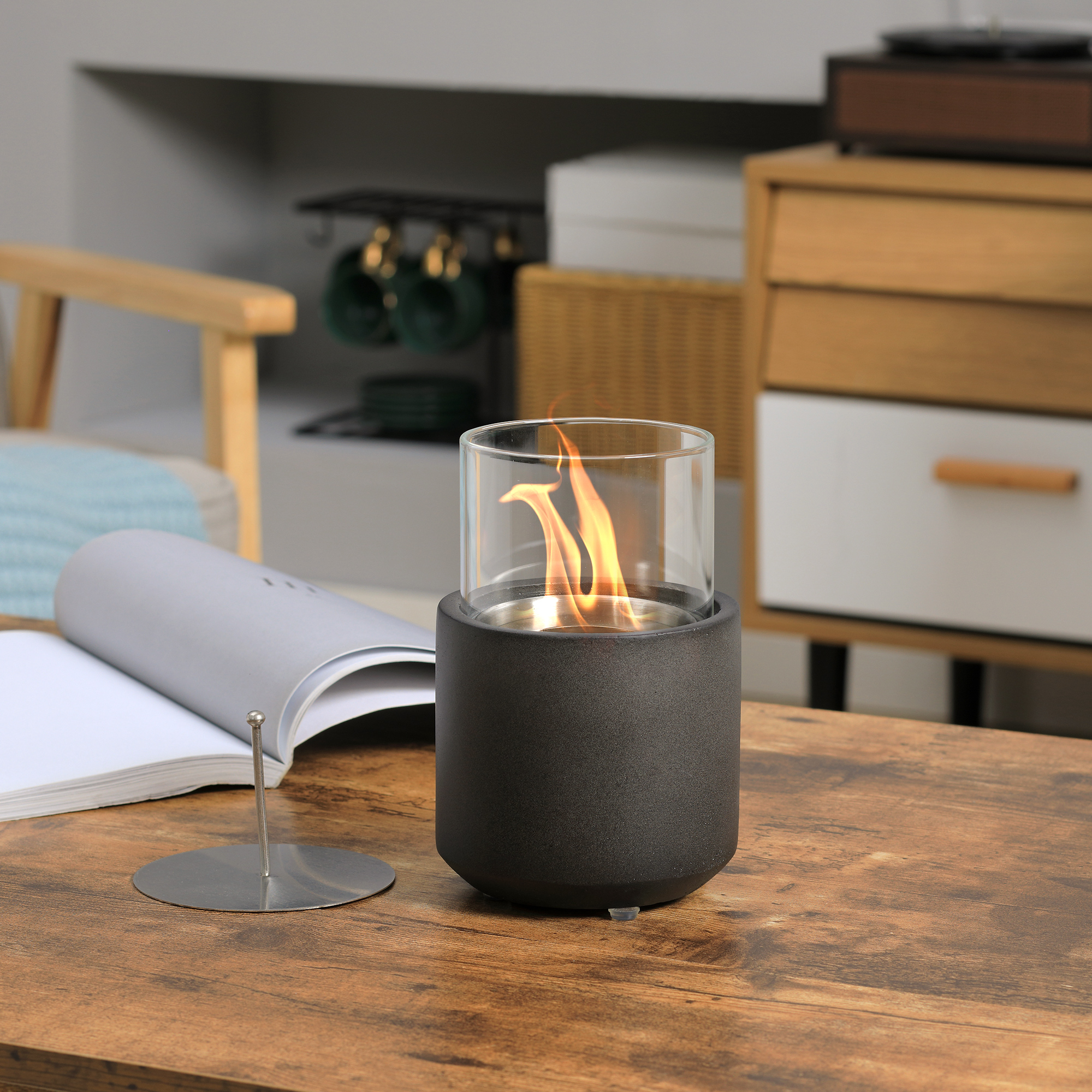 Un caminetto cilindrico nero acceso su un tavolo di legno, con libro aperto e decorazioni.