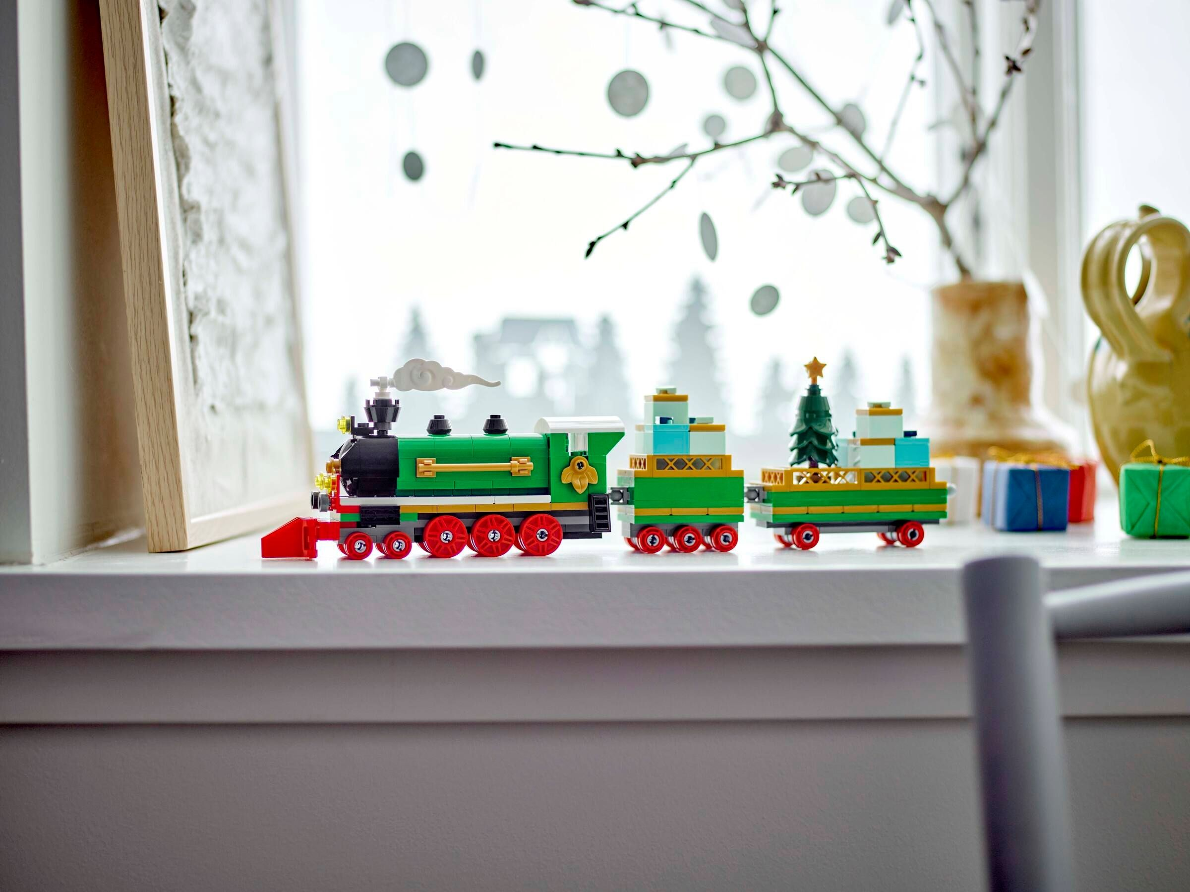 Ein Spielzeug-Zug-Set auf einer Fensterbank. Der Zug ist grün, rot und gold, mit einem Weihnachtsbaum auf einem Wagen.