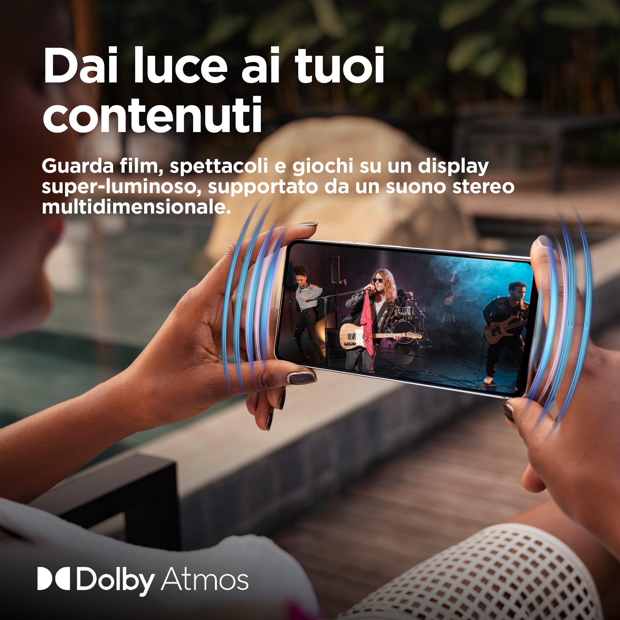 Osoba trzymająca telefon z zespołem na żywo. Niebieskie fale dźwiękowe promieniują z telefonu. Logo Dolby Atmos.