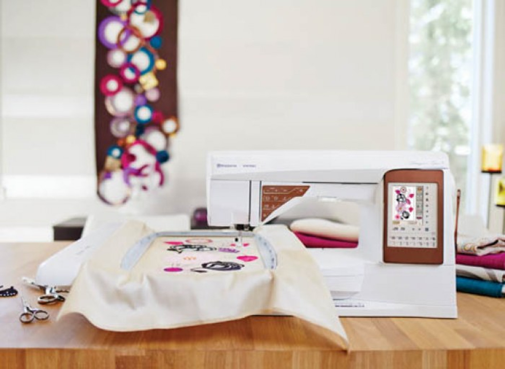 Máquina de coser con bordado. Pantalla muestra diseño, tela asegurada en aro. Mesa y fondo.