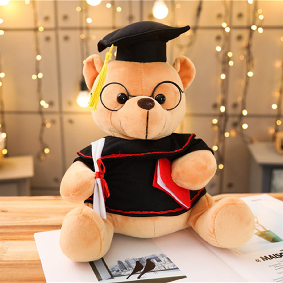Teddybär mit Abschlusskappe, Brille und Robe. Hält ein Diplom und ein rotes Buch. Sitzt auf einem Buch.