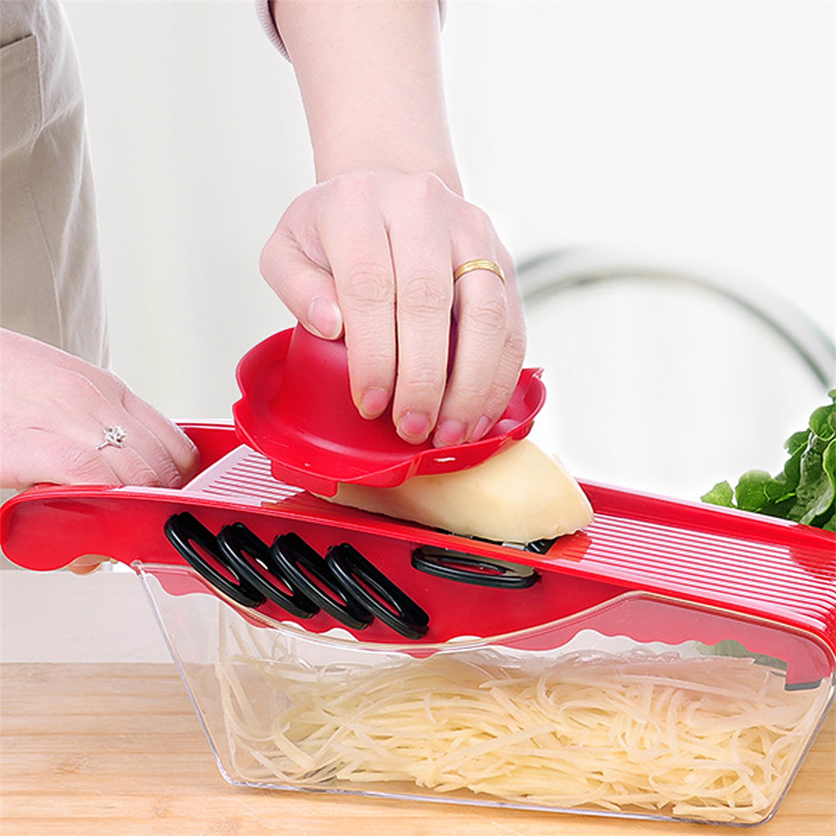 Eine Person benutzt eine rote Mandoline mit Sicherheitsgriff, um eine Kartoffel zu schneiden. Geschnittene Kartoffeln sammeln sich darunter.