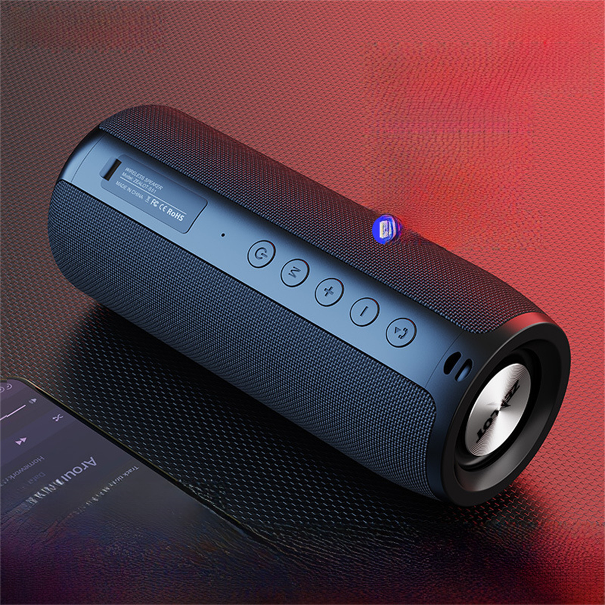 Ein schwarzer zylindrischer Lautsprecher mit Knöpfen, einem Licht und einem Handy mit Musik-App auf roter Fläche.