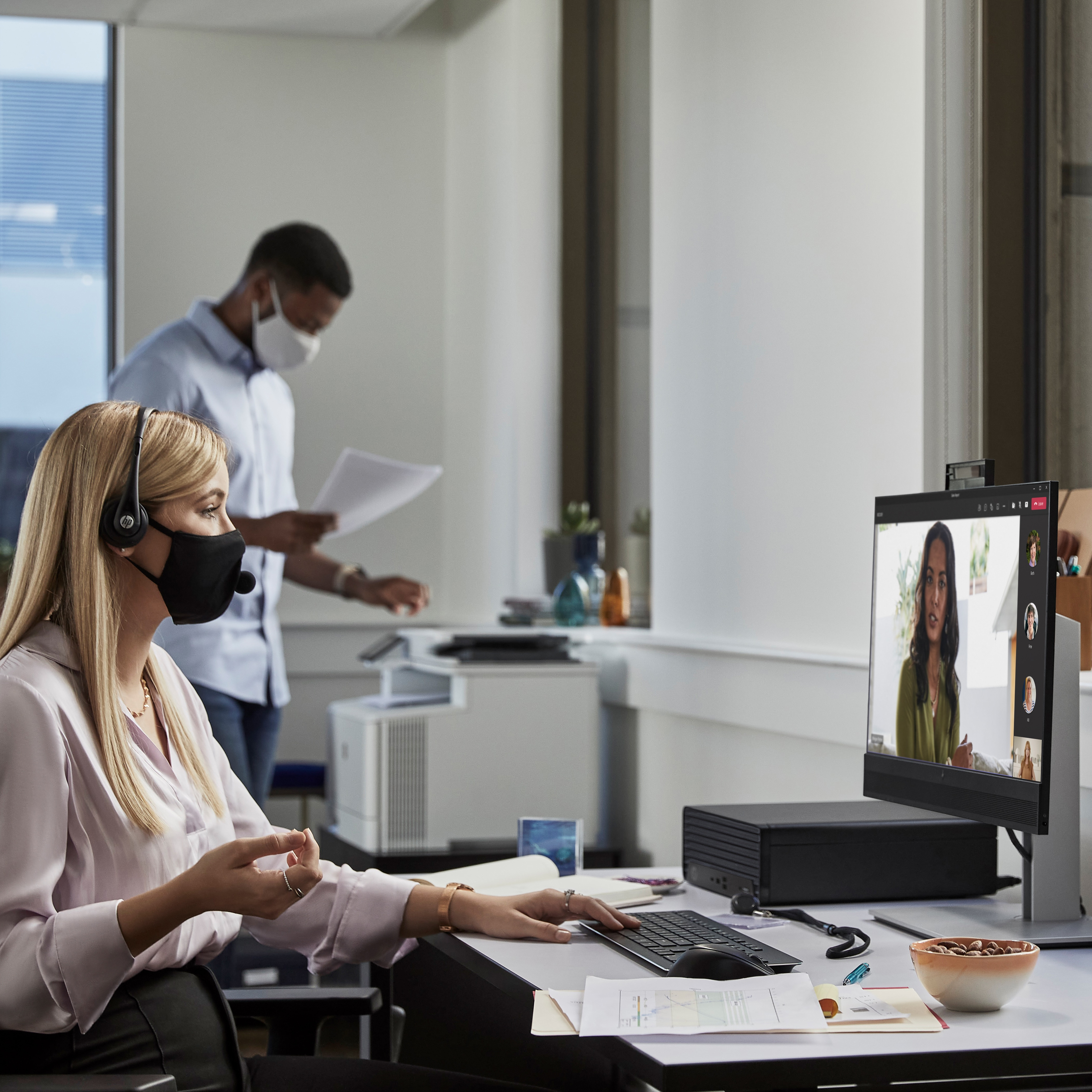 Scena biurowa: kobieta z zestawem słuchawkowym na rozmowie wideo, mężczyzna w tle, oboje w maskach.