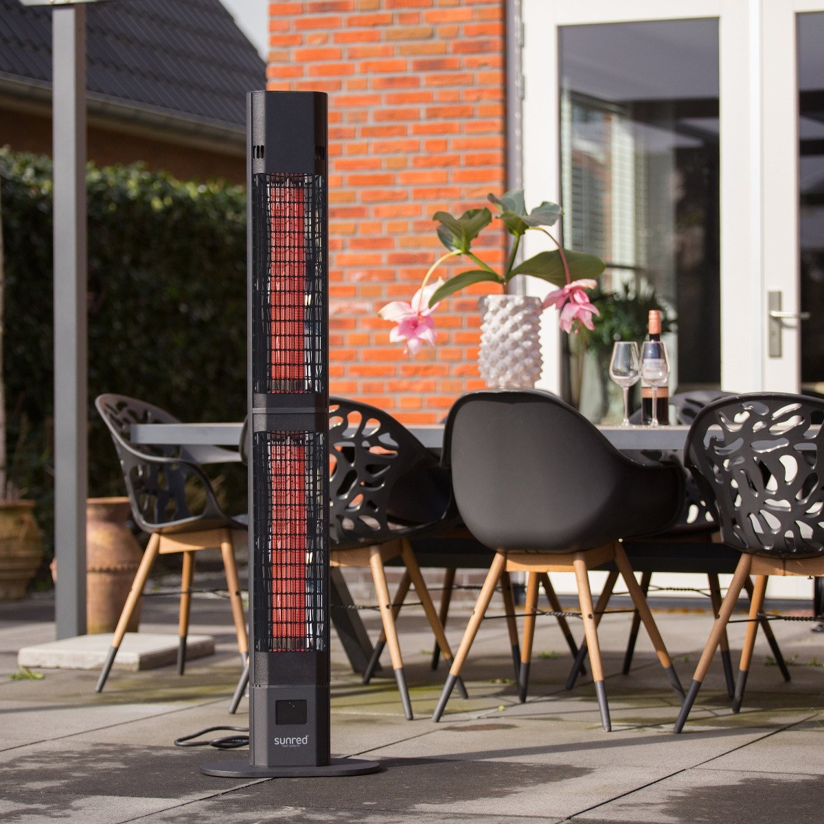 An outdoor patio scene with a black heater, table, chairs, and plants, with a red brick wall.
