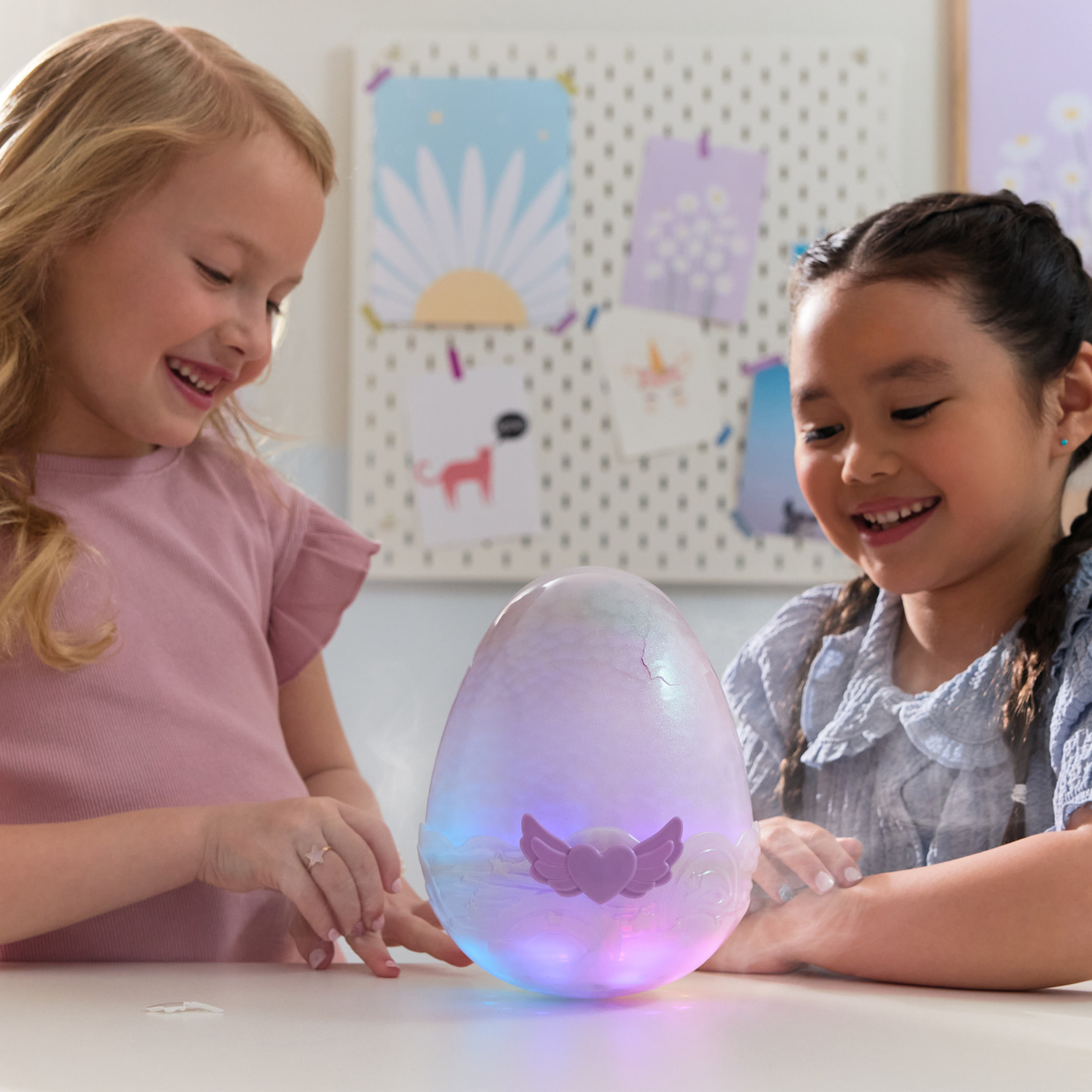 Dos niñas sonriendo, mirando un juguete en forma de huevo que brilla. El juguete tiene alas y se ilumina con varios colores.