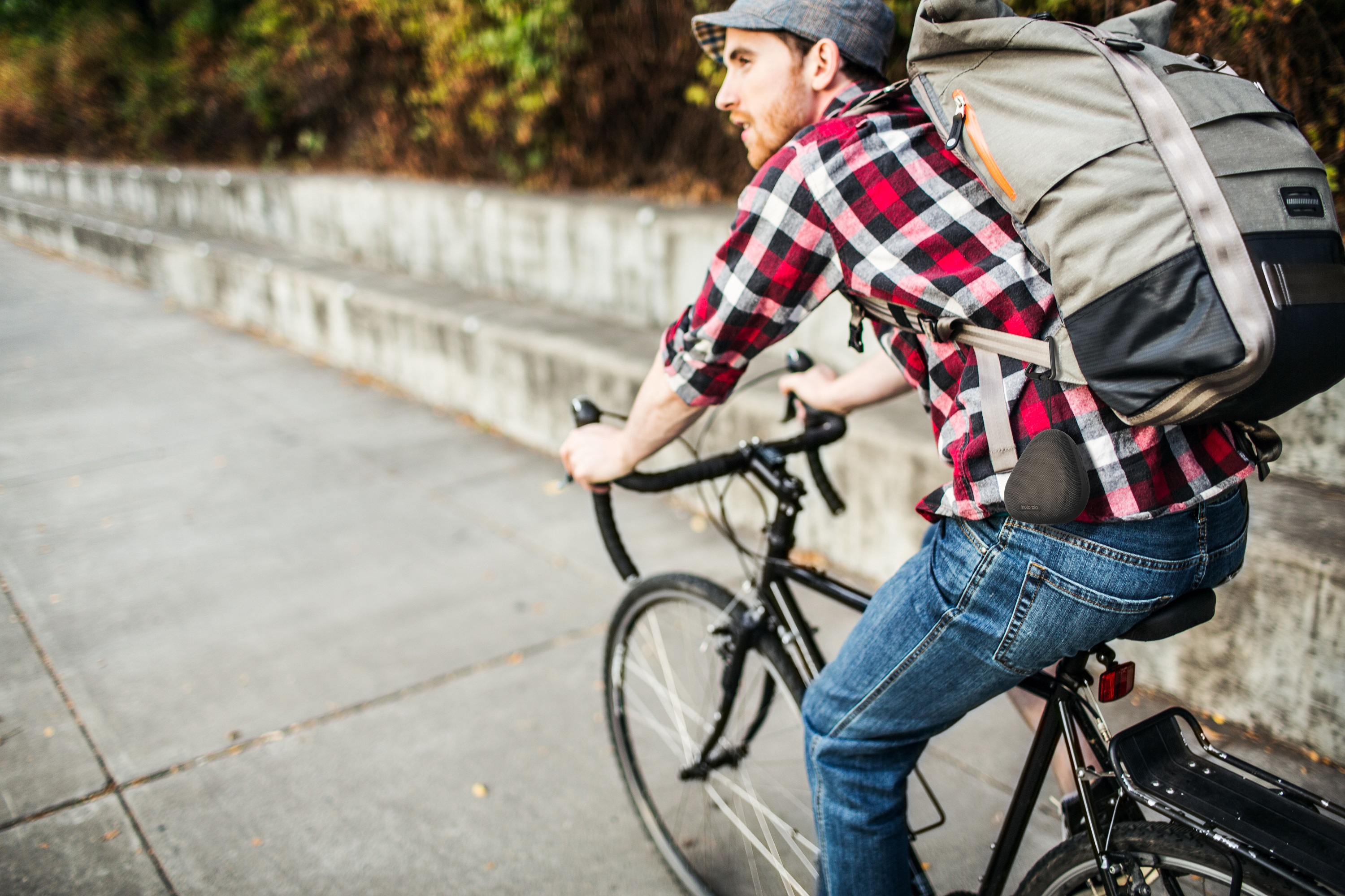 Hombre montando en bicicleta en una acera. Viste una camisa a cuadros, jeans y una gorra, con una mochila.