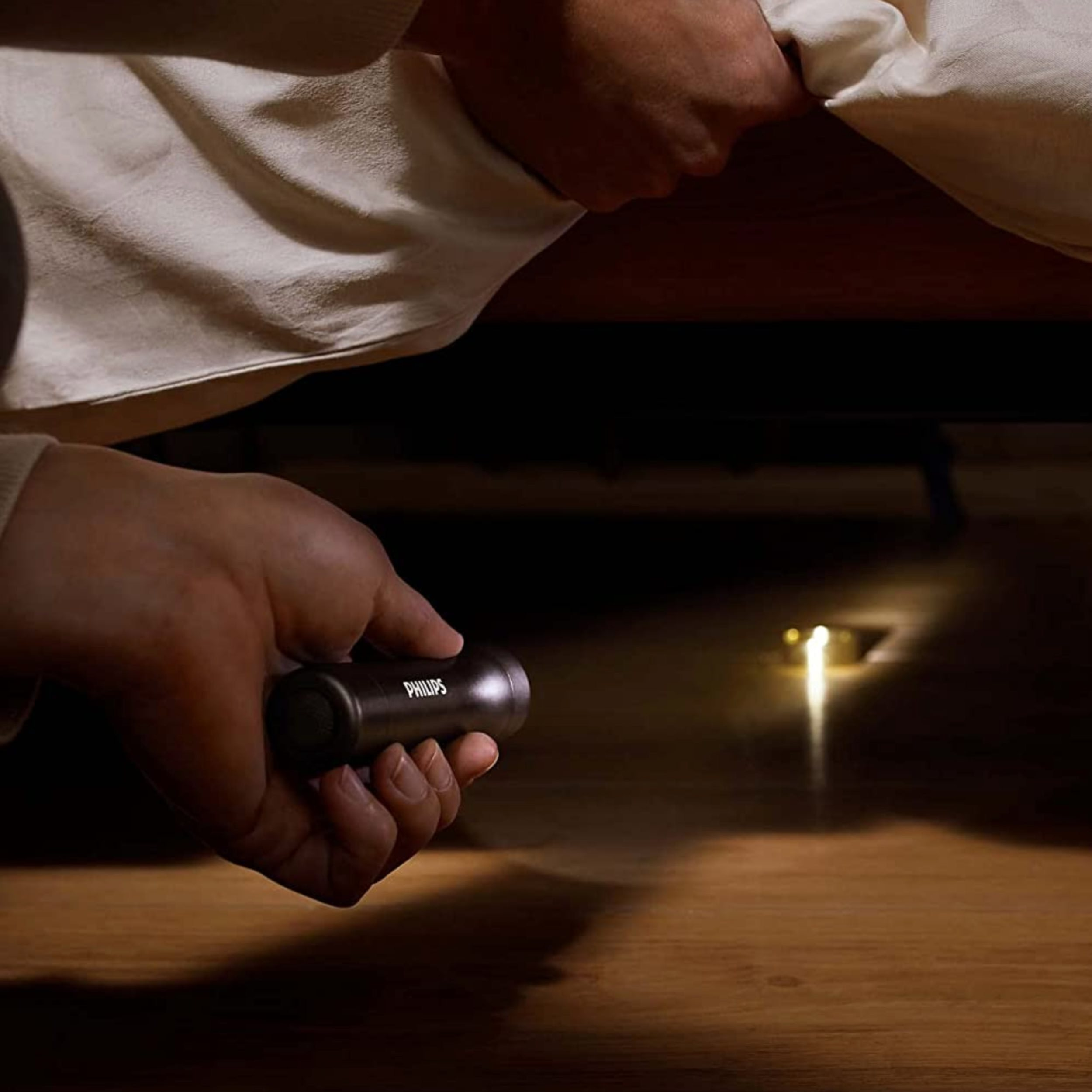 Una mano che tiene una torcia nera, che illumina una superficie di legno scuro, vicino a un letto.