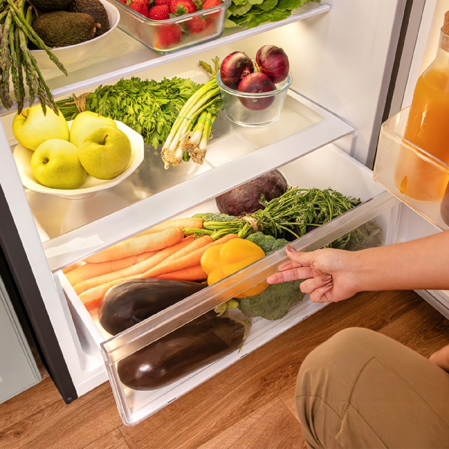 Wnętrze lodówki z warzywami. Ręka wyciąga szufladę z marchewką, bakłażanem i papryką.