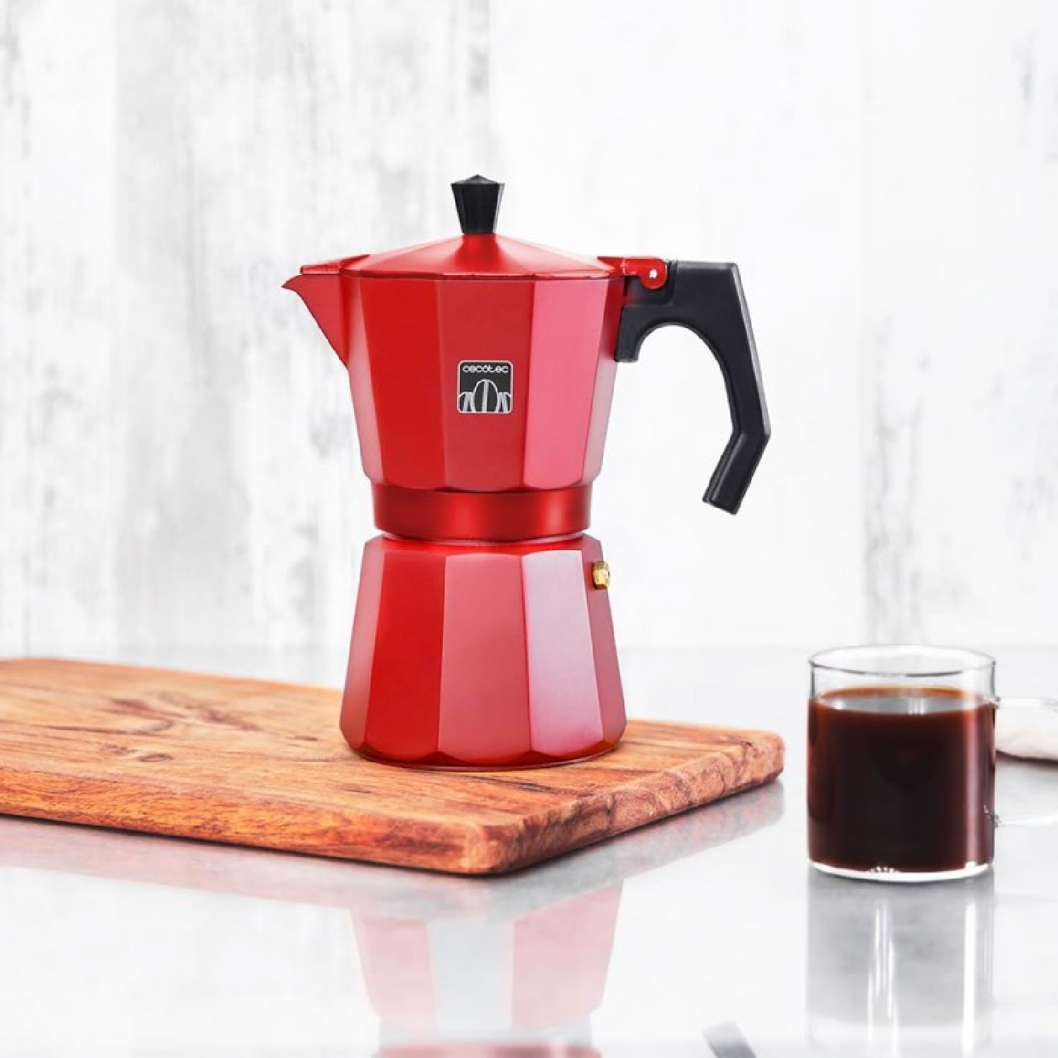 Roter Espressokocher und ein Glas Kaffee auf einem Holzbrett, auf weißer Marmoroberfläche.