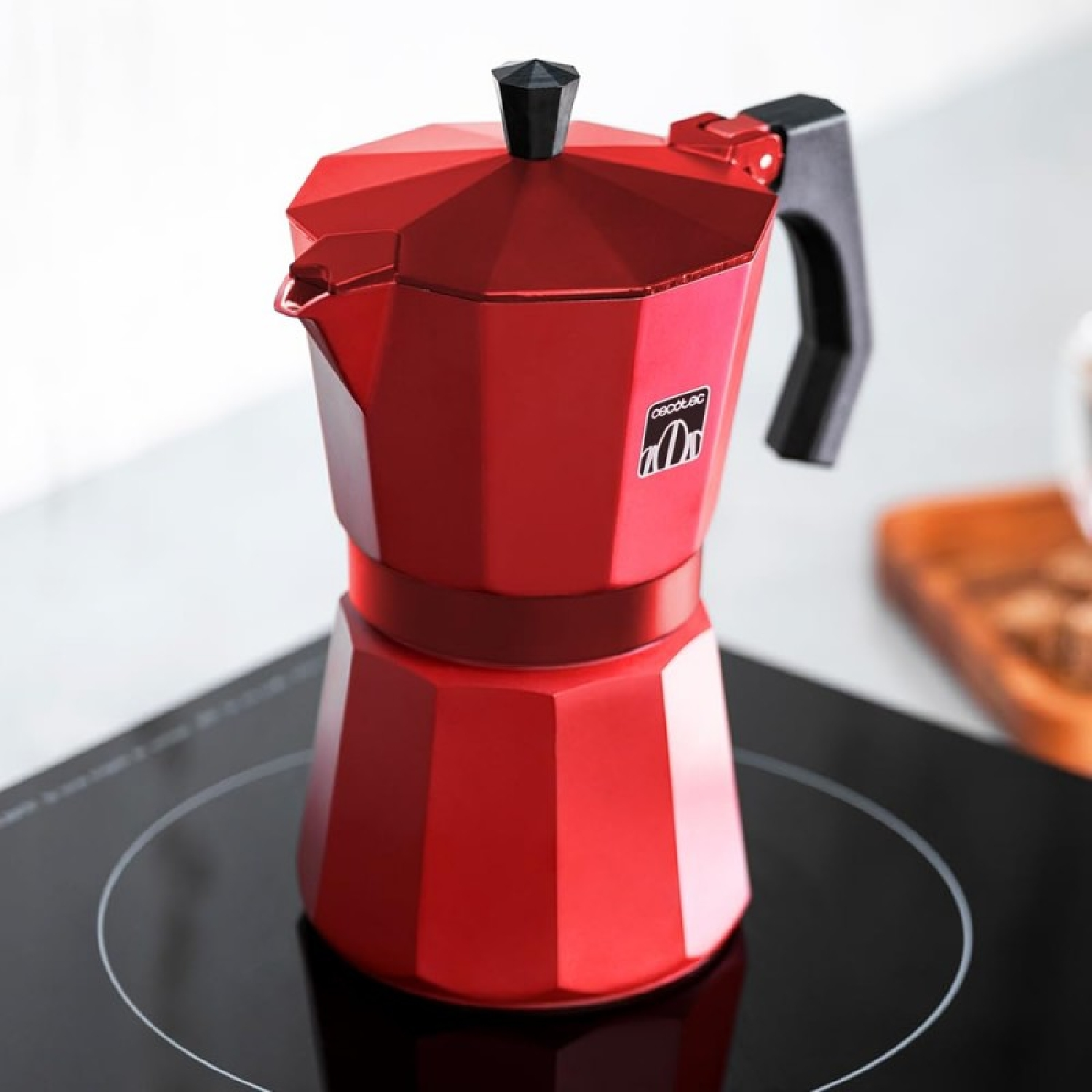 Roter Kaffeezubereiter auf einem schwarzen Kochfeld, schräge Ansicht. Der Bereiter hat einen schwarzen Griff und Deckel.