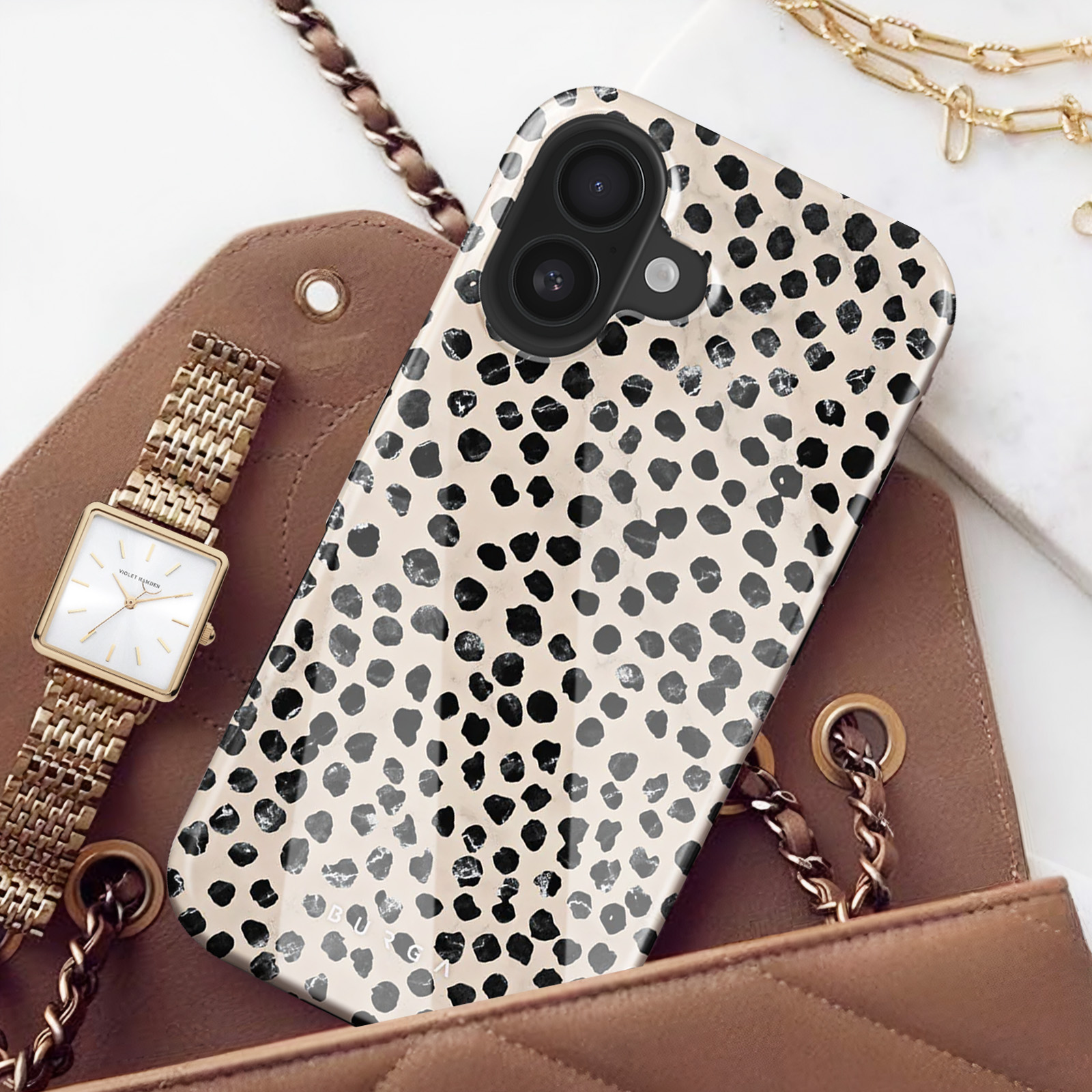 Een telefoonhoesje met zwarte stippen op een beige achtergrond. Items zoals een horloge en een tas liggen op tafel.