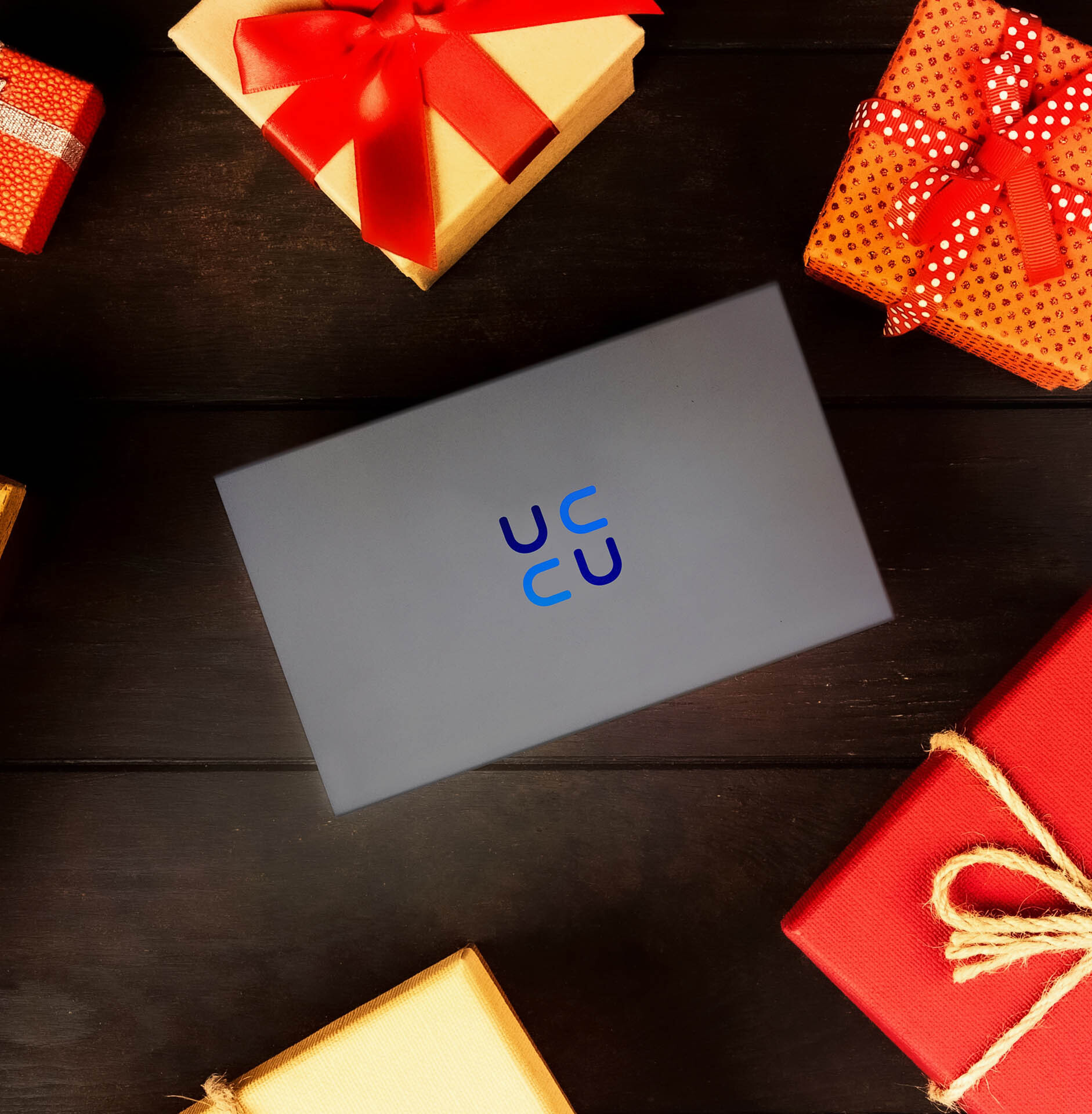 Eine graue Box mit Firmenlogo, umgeben von Geschenken in verschiedenen Farben, alles auf dunkler Holzoberfläche.