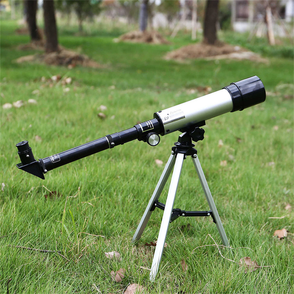 Ein silbernes Teleskop auf einem Stativ, auf grünem Gras mit Bäumen im Hintergrund.