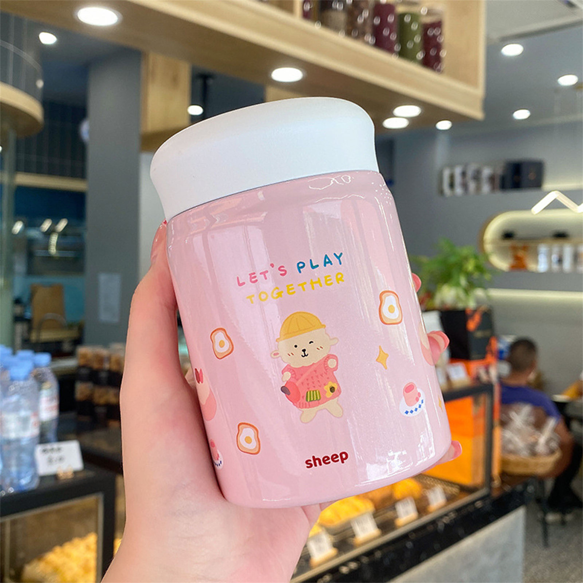 Ein rosa Becher mit weißem Deckel, mit einer Cartoon-Schaf und den Worten 'LET'S PLAY TOGETHER'.